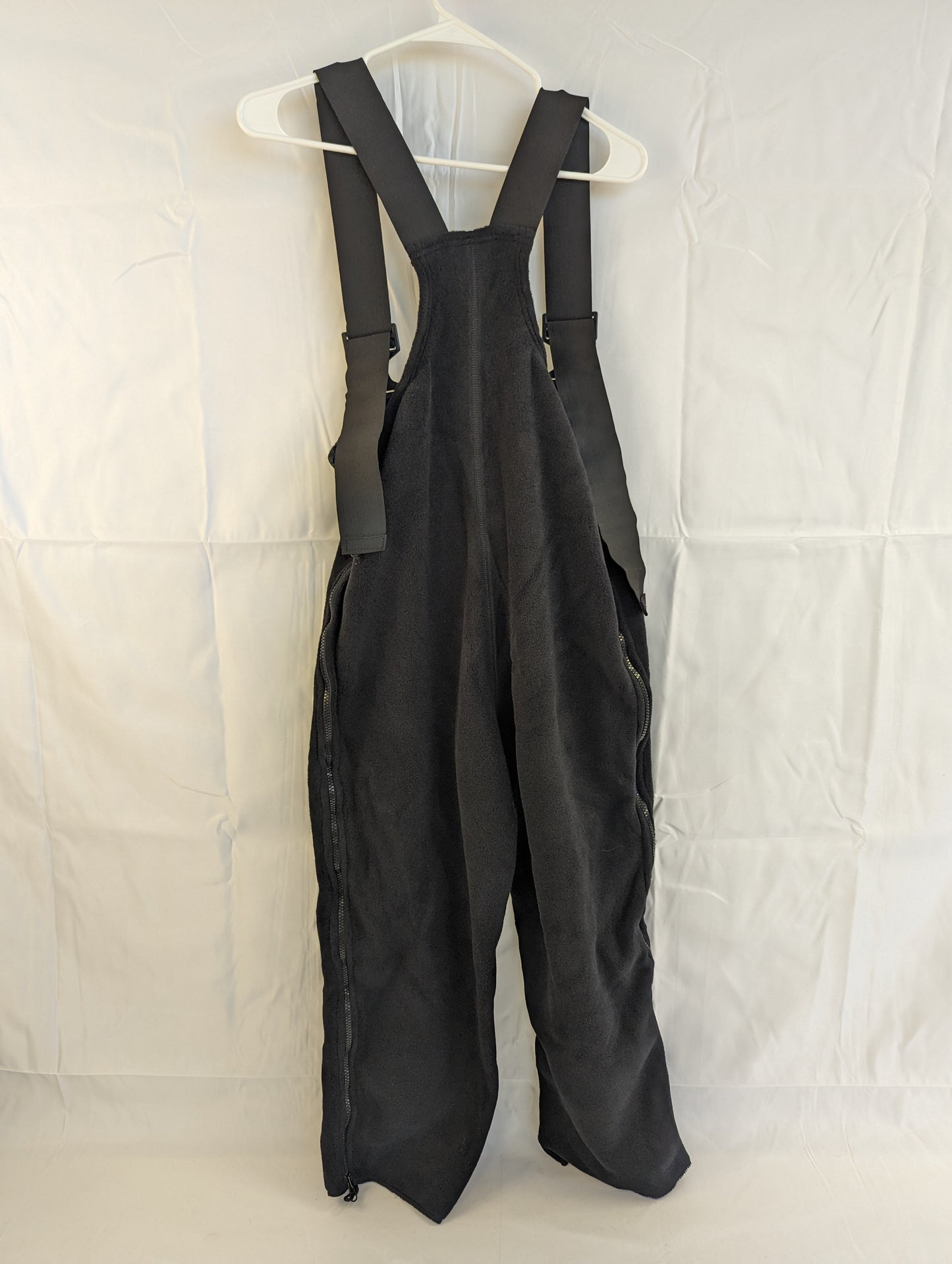 A full backview of black Polartec fleece cold weather overalls hanging on a hanger, featuring adjustable shoulder straps with buckles and full-length side zippers.