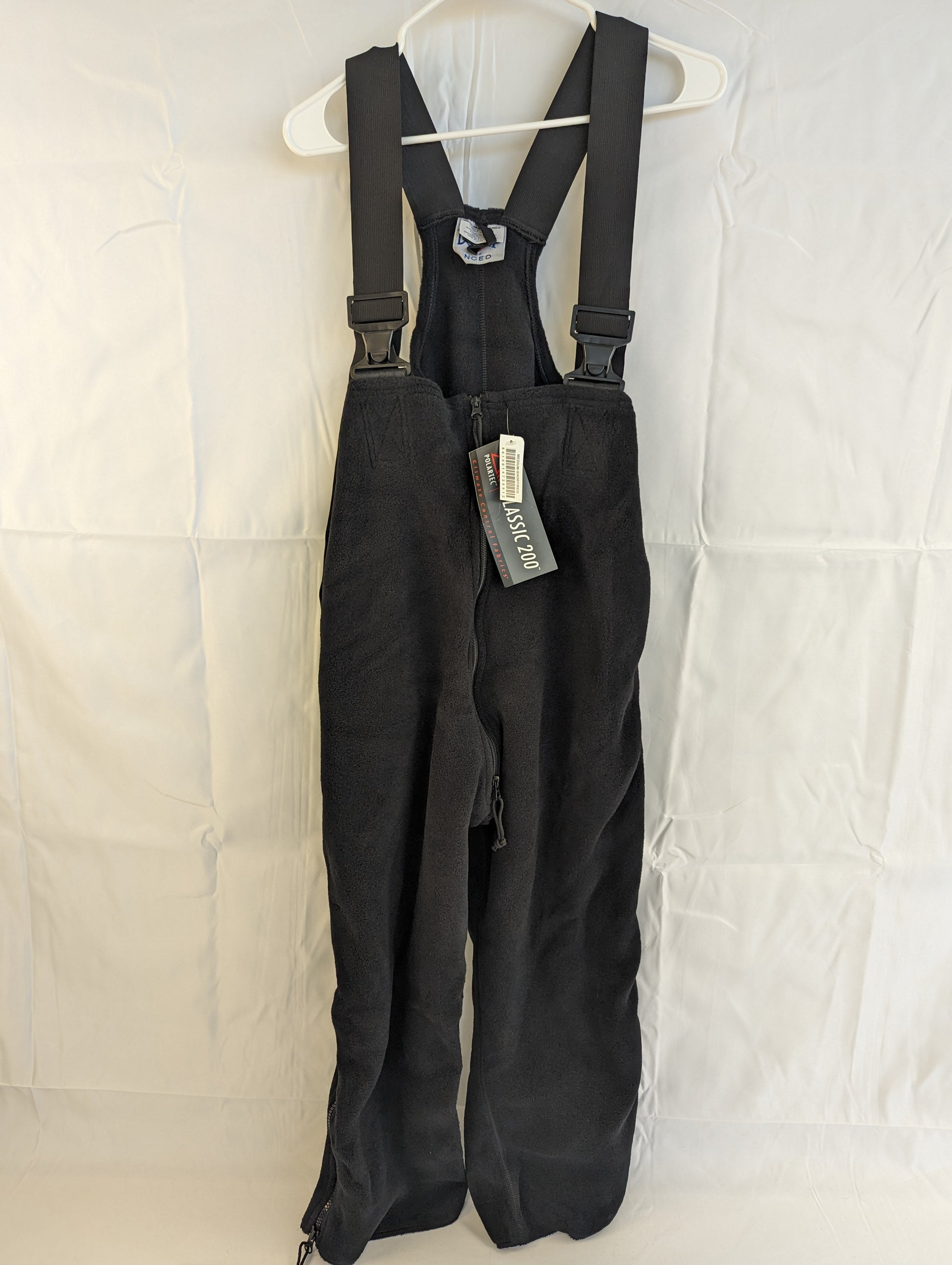 A full front view of black Polartec fleece cold weather overalls hanging on a hanger, featuring adjustable shoulder straps with buckles and the original tag attached.