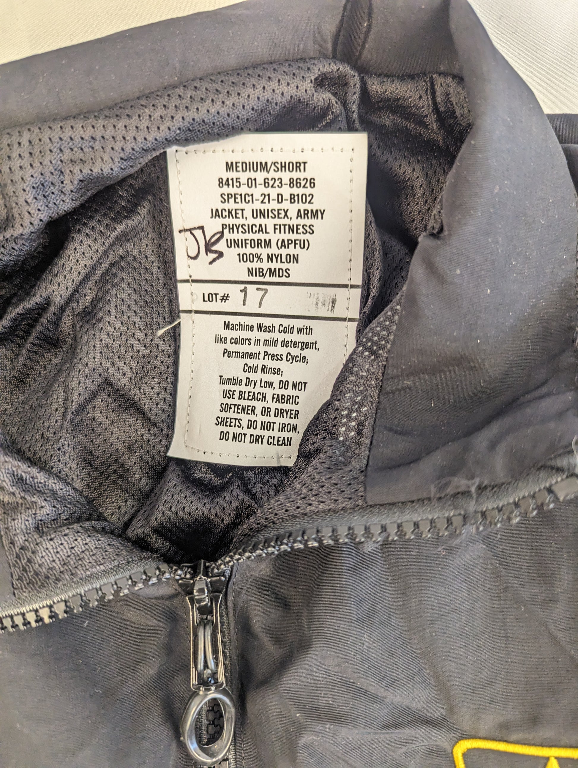 A close-up photograph of the U.S. Army Physical Fitness Uniform (APFU) jacket issue tag, clearly displaying the size 'MEDIUM/SHORT', the National Stock Number (NSN) 8415-01-623-8626, and material details.
