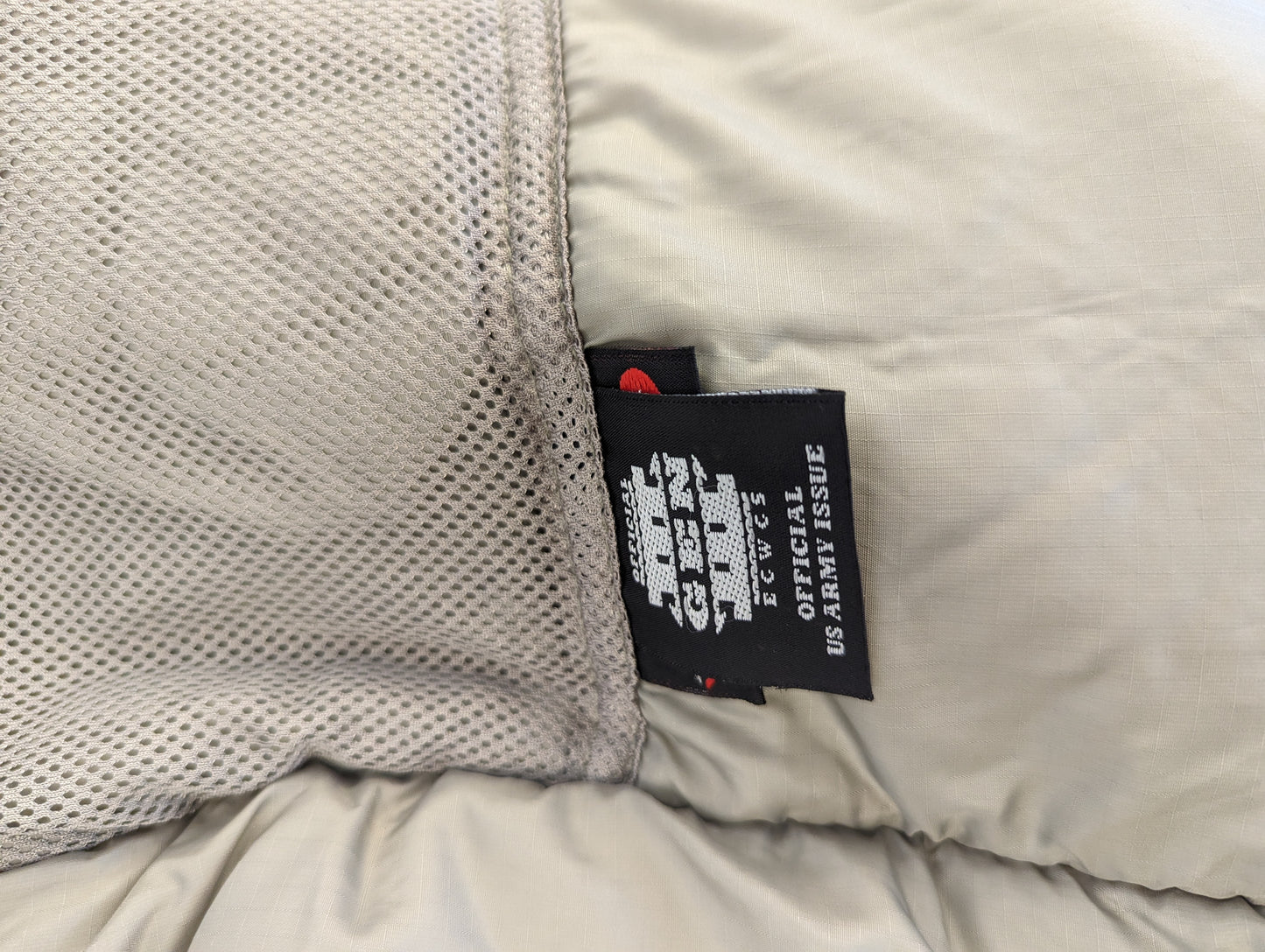 A close-up photograph of the black official U.S. Army issue tag sewn into the Extreme Cold Weather Parka, prominently displaying 'ECWCS GEN III', 'OFFICIAL U.S. ARMY ISSUE', and 'Made in U.S.A.' branding.