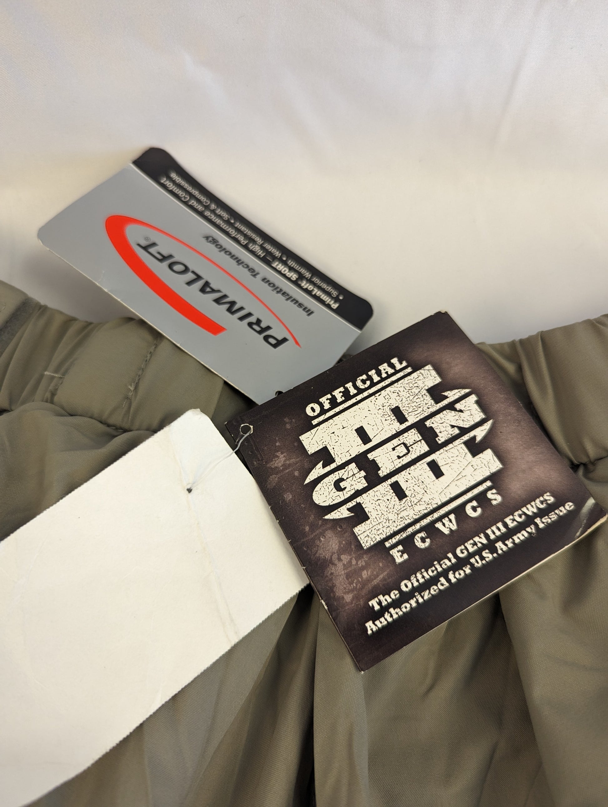 A close-up photograph of the official tags sewn into the U.S. Military Extreme Cold Weather Trousers, displaying the 'PRIMALOFT' insulation logo and the 'OFFICIAL U.S. ARMY ISSUE' GEN III ECWCS label.