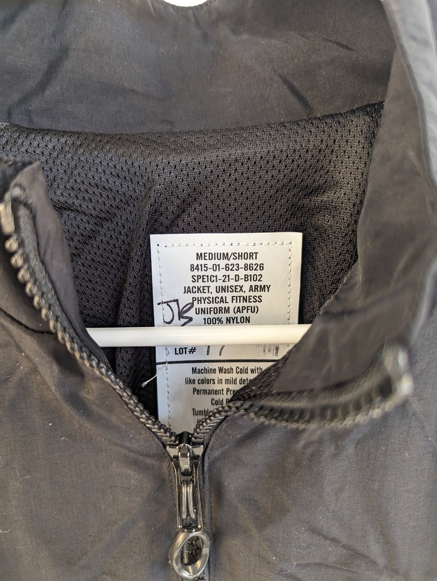 A close-up photograph of the U.S. Army Physical Fitness Uniform (APFU) jacket issue tag, clearly displaying the size 'MEDIUM/SHORT', the National Stock Number (NSN), and material details.