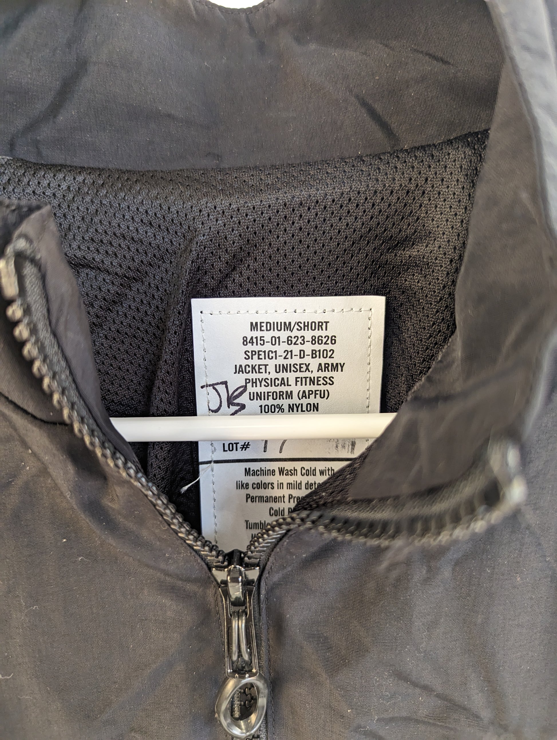 A close-up photograph of the U.S. Army Physical Fitness Uniform (APFU) jacket issue tag, clearly displaying the size 'MEDIUM/SHORT', the National Stock Number (NSN), and material details.