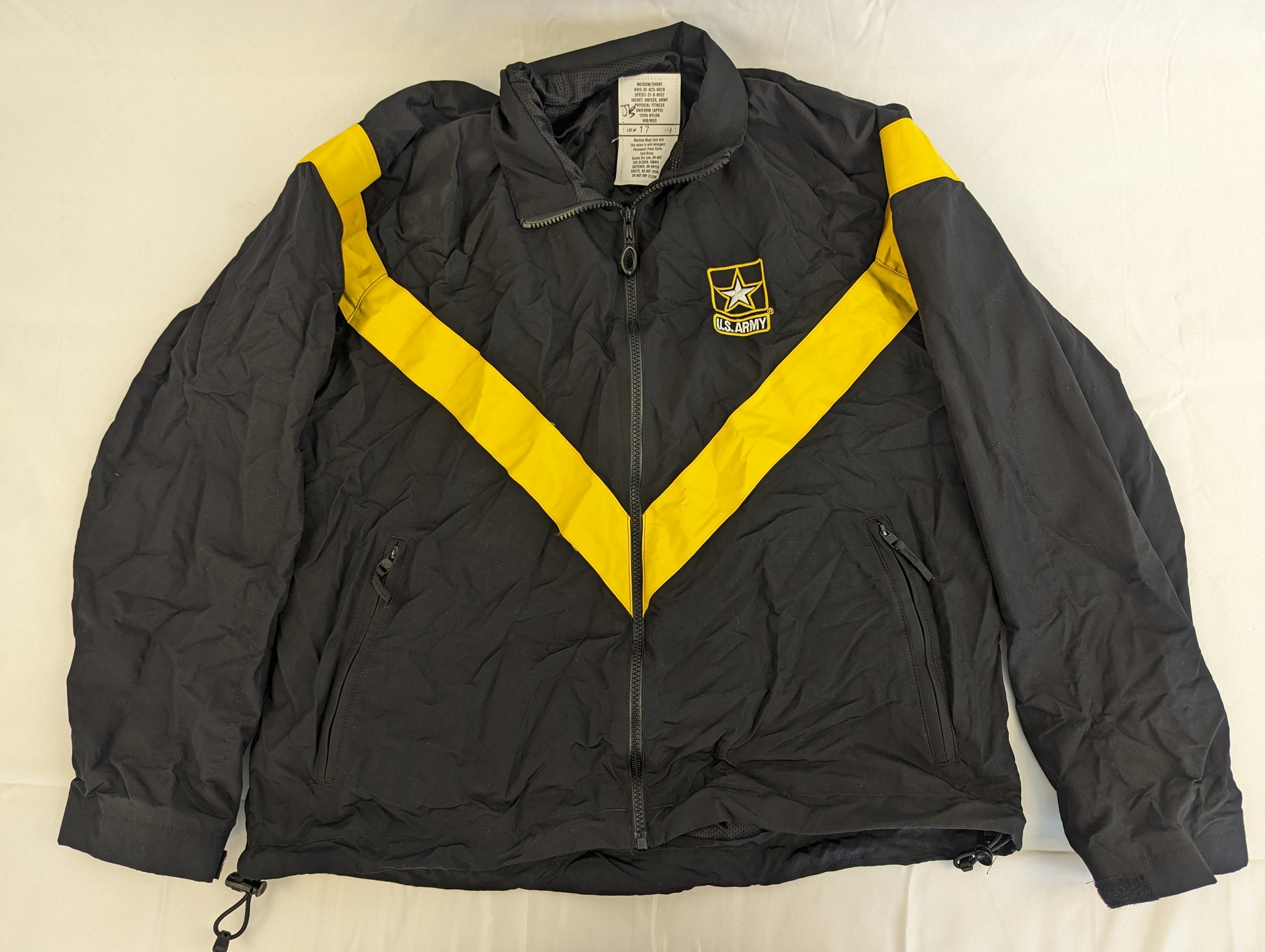A front view of a black U.S. Army Physical Training (PT) jacket laid flat on a white background, featuring a bold yellow chevron stripe across the chest and the embroidered U.S. Army logo on the left breast.