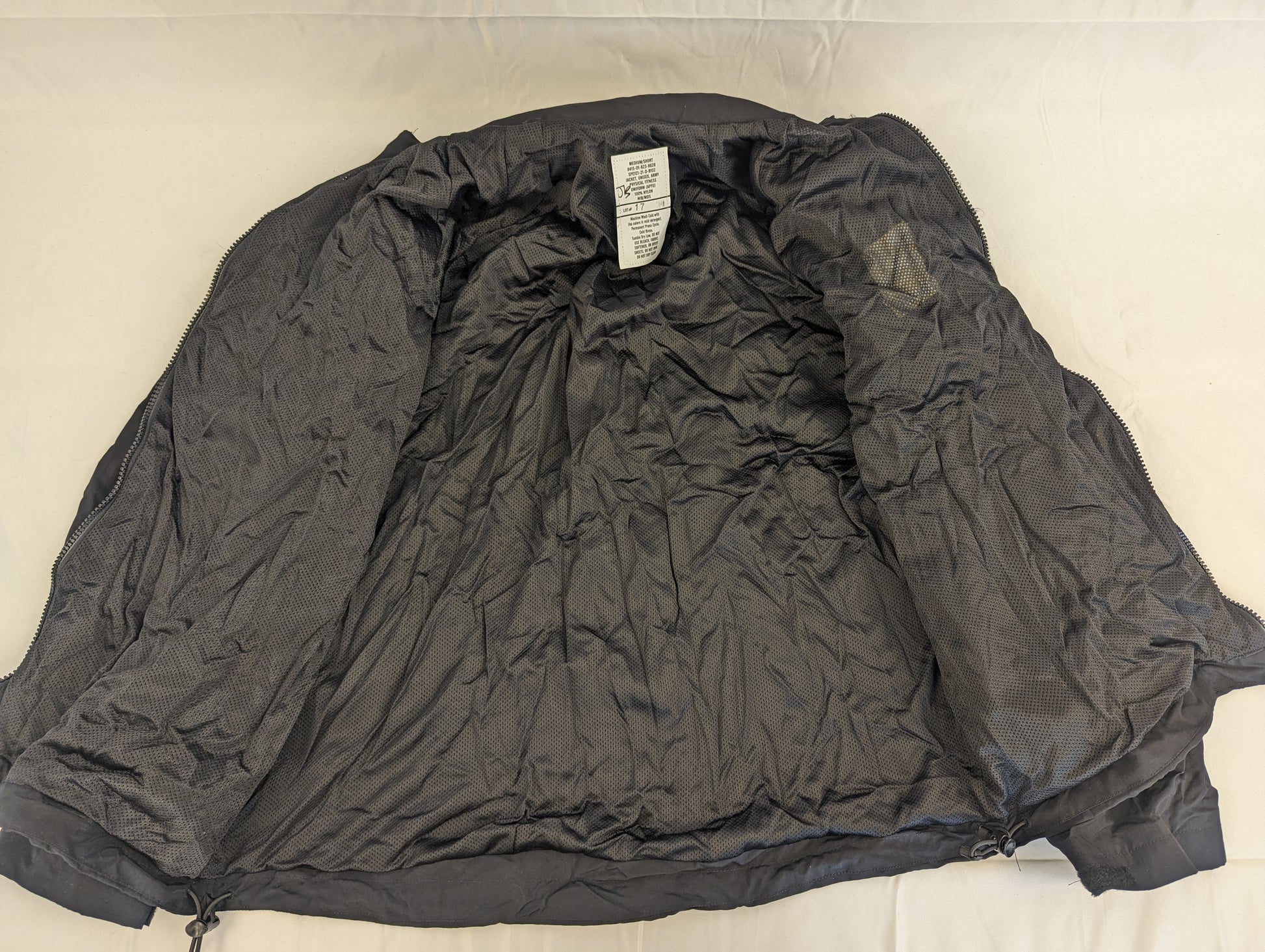 An open view of the black U.S. Army Physical Training (PT) jacket laid flat, displaying the breathable black mesh lining and the size tag at the collar.