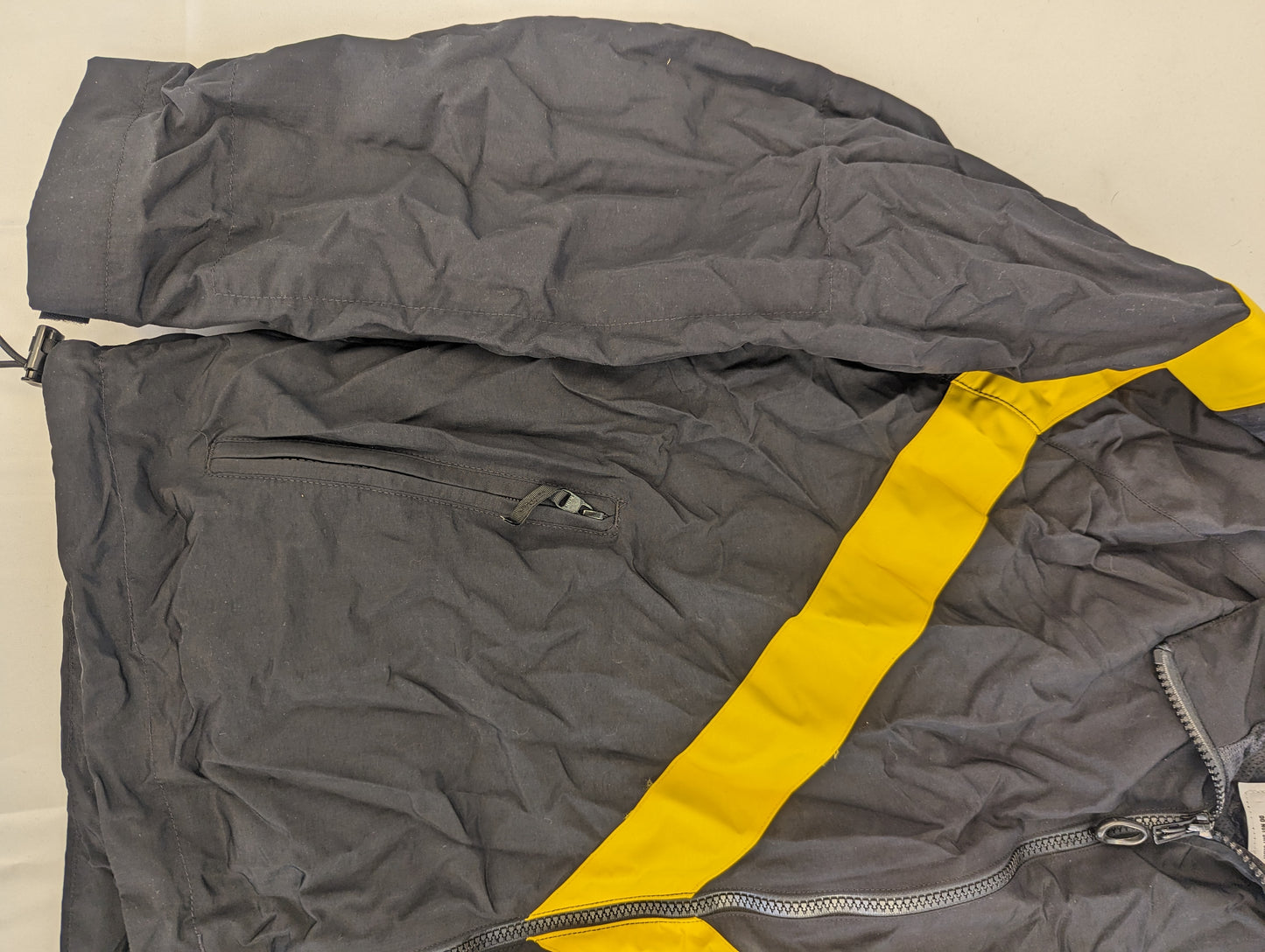 A close-up view of the black U.S. Army Physical Training (PT) jacket, highlighting the diagonal yellow chevron stripe, the zippered hand pocket, and the elastic sleeve cuff.