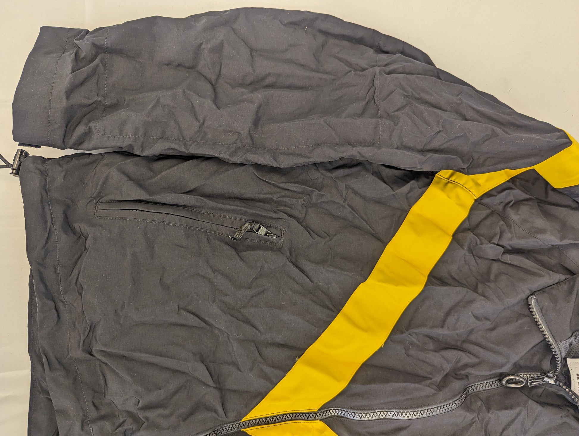 A close-up view of the black U.S. Army Physical Training (PT) jacket, highlighting the diagonal yellow chevron stripe, the zippered hand pocket, and the elastic sleeve cuff.