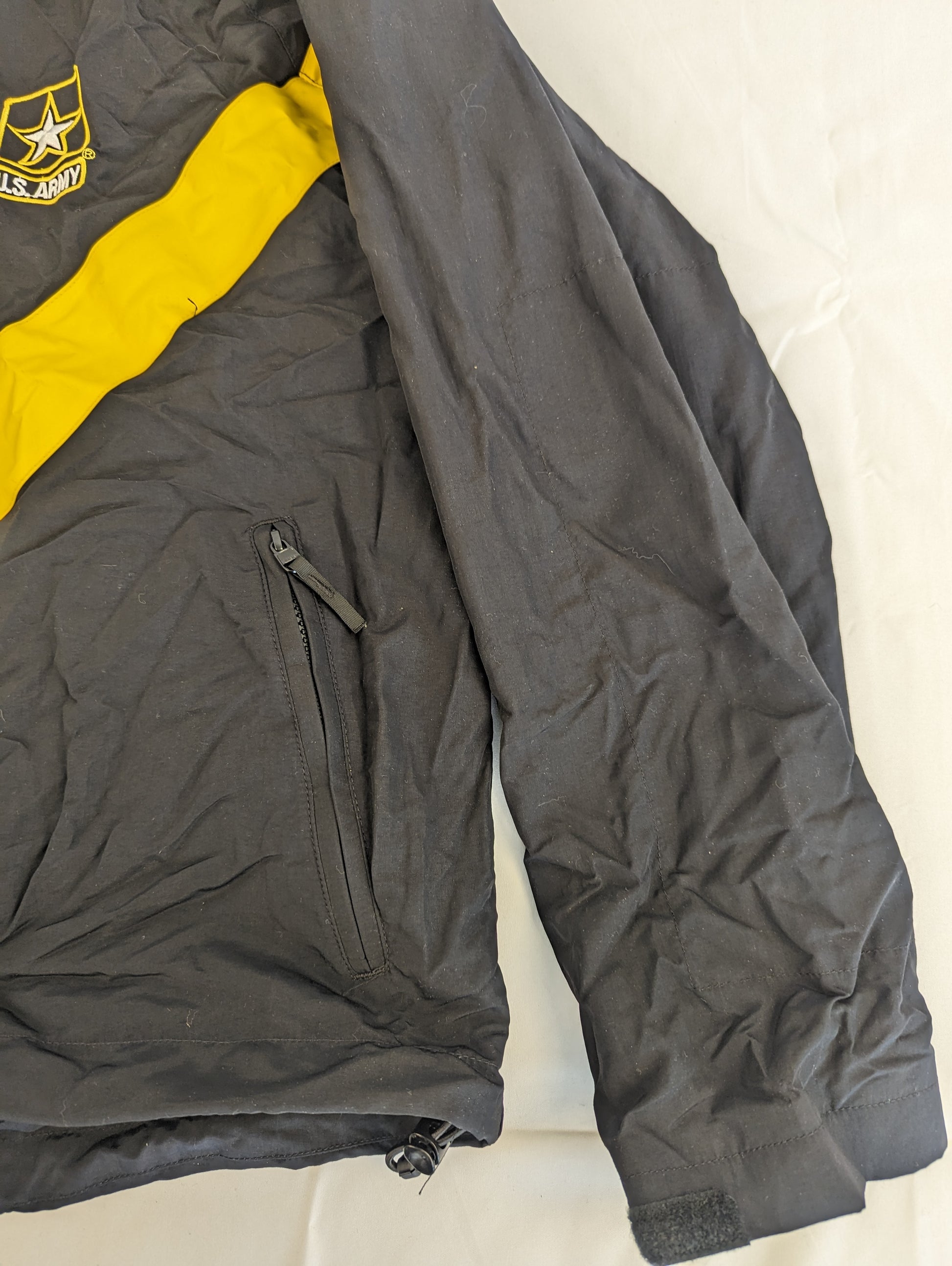 A close-up view of the side and sleeve of a black U.S. Army Physical Training (PT) jacket, highlighting the zippered hand pocket, the sleeve cuff, and the yellow chevron stripe on the chest.