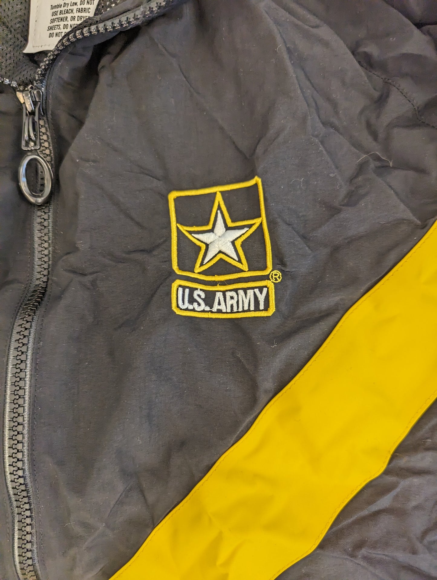 A close-up detail shot of the black U.S. Army Physical Training (PT) jacket, showing the embroidered U.S. Army star logo, the diagonal yellow chevron stripe, and the front zipper.