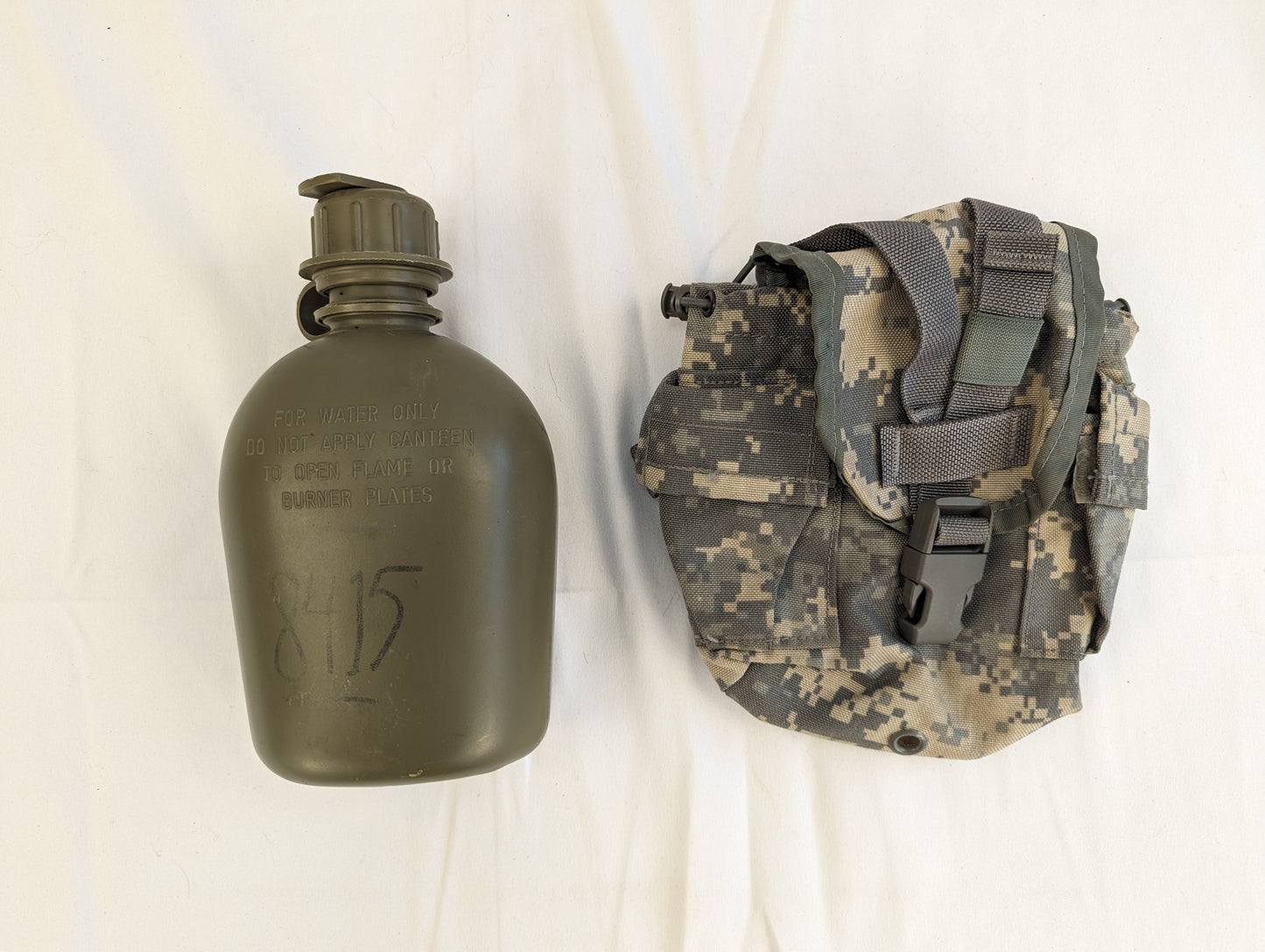 A used U.S. Military Surplus 1 Quart Water Canteen (olive green with a black marker serial number) shown next to its matching ACU digital camouflage MOLLE pouch, resting on a white background.