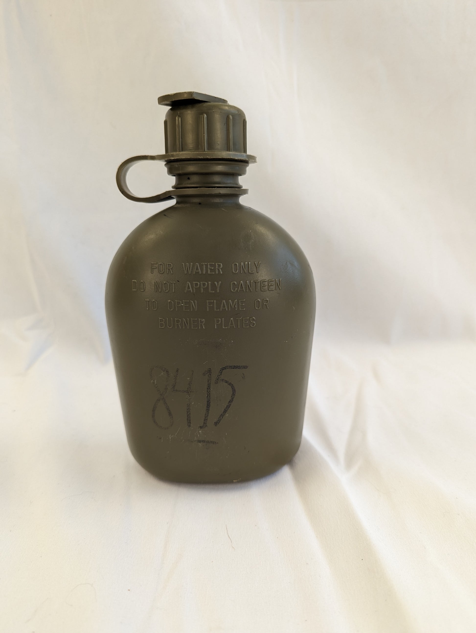 A close-up studio shot of a used U.S. Military Surplus 1-quart plastic canteen in olive green with a black cap, showing faint molded text and a visible handwritten military stock number.