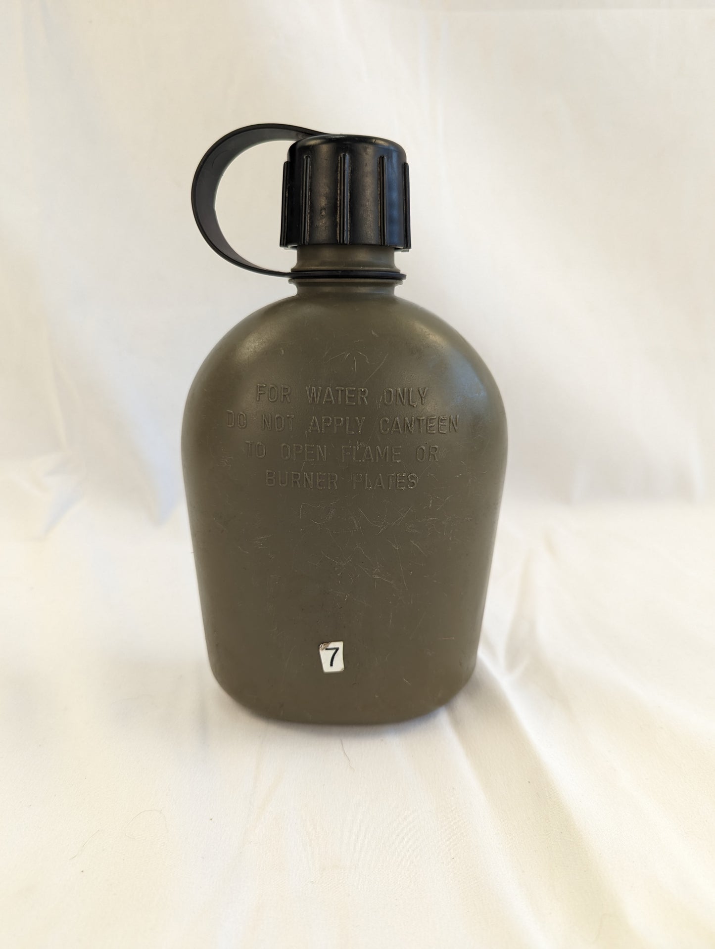 A close-up studio shot of a used U.S. Military Surplus 1-quart plastic canteen in olive green with a black cap, standing upright on a wrinkled white cloth background.