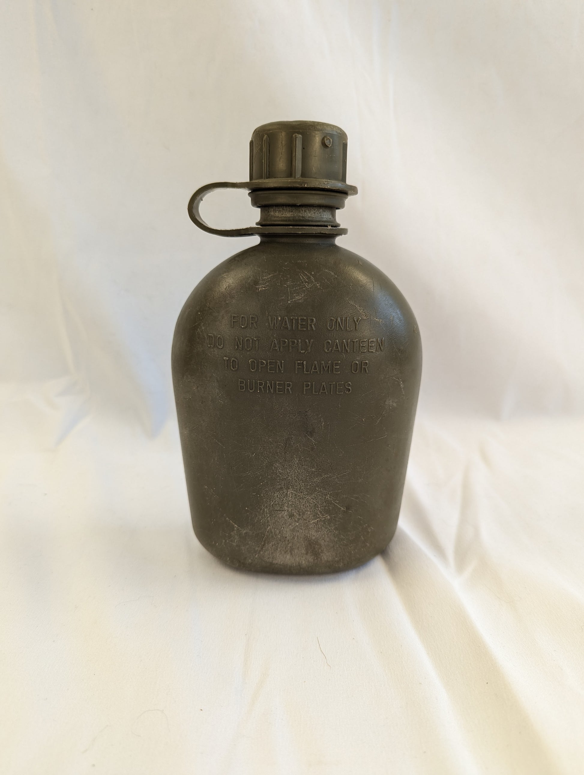 A close-up studio shot of a used U.S. Military Surplus 1-quart plastic canteen in olive green, standing upright on a wrinkled white cloth background.