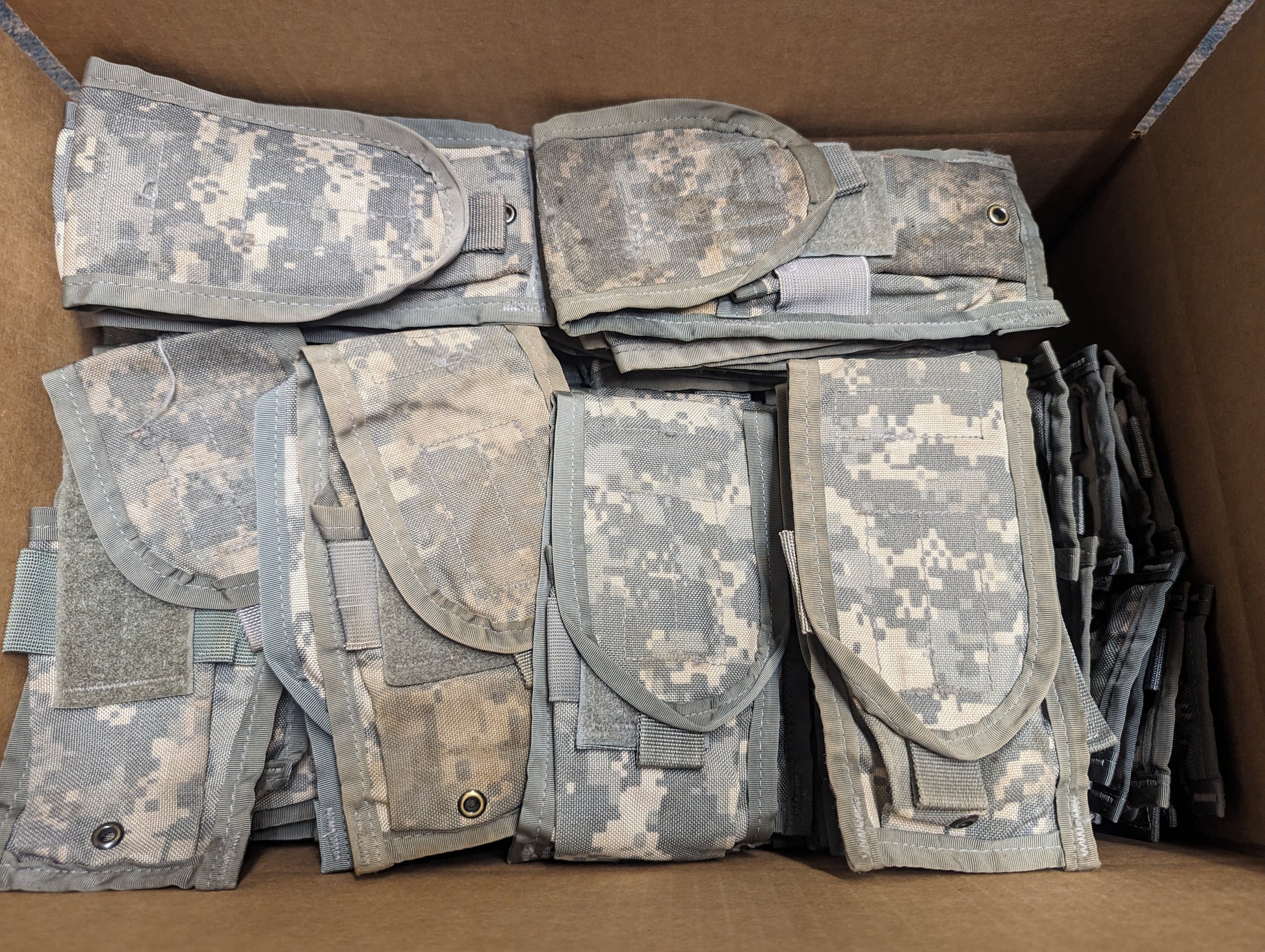 A cardboard box filled with approximately one hundred stacked and folded U.S. Military Surplus 2-Mag Pouches in ACU digital camouflage.