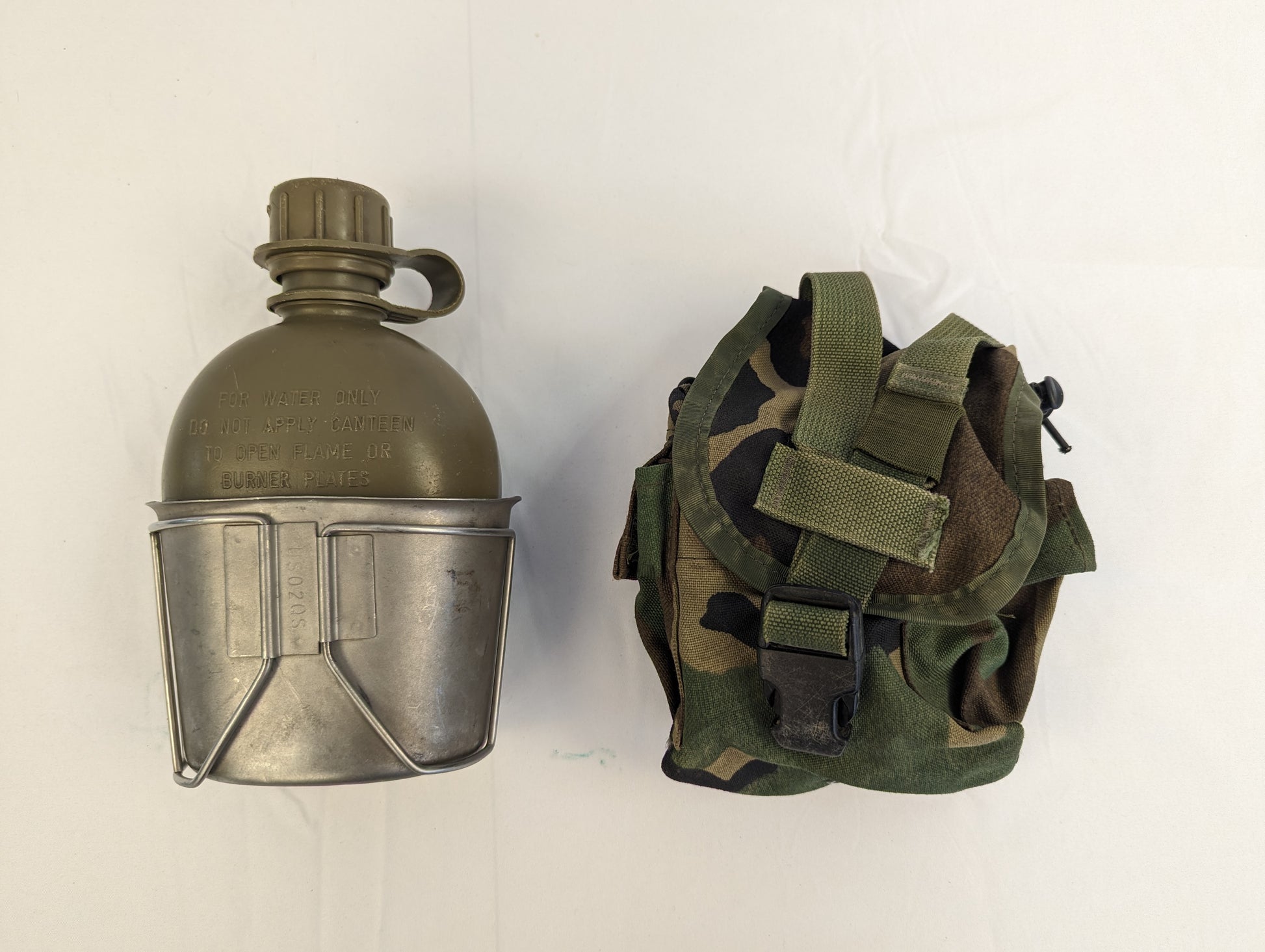 A studio shot of a U.S. Military 1-quart canteen set, featuring an olive green plastic canteen nested inside a metal cup, displayed next to a closed Woodland camouflage fabric pouch.