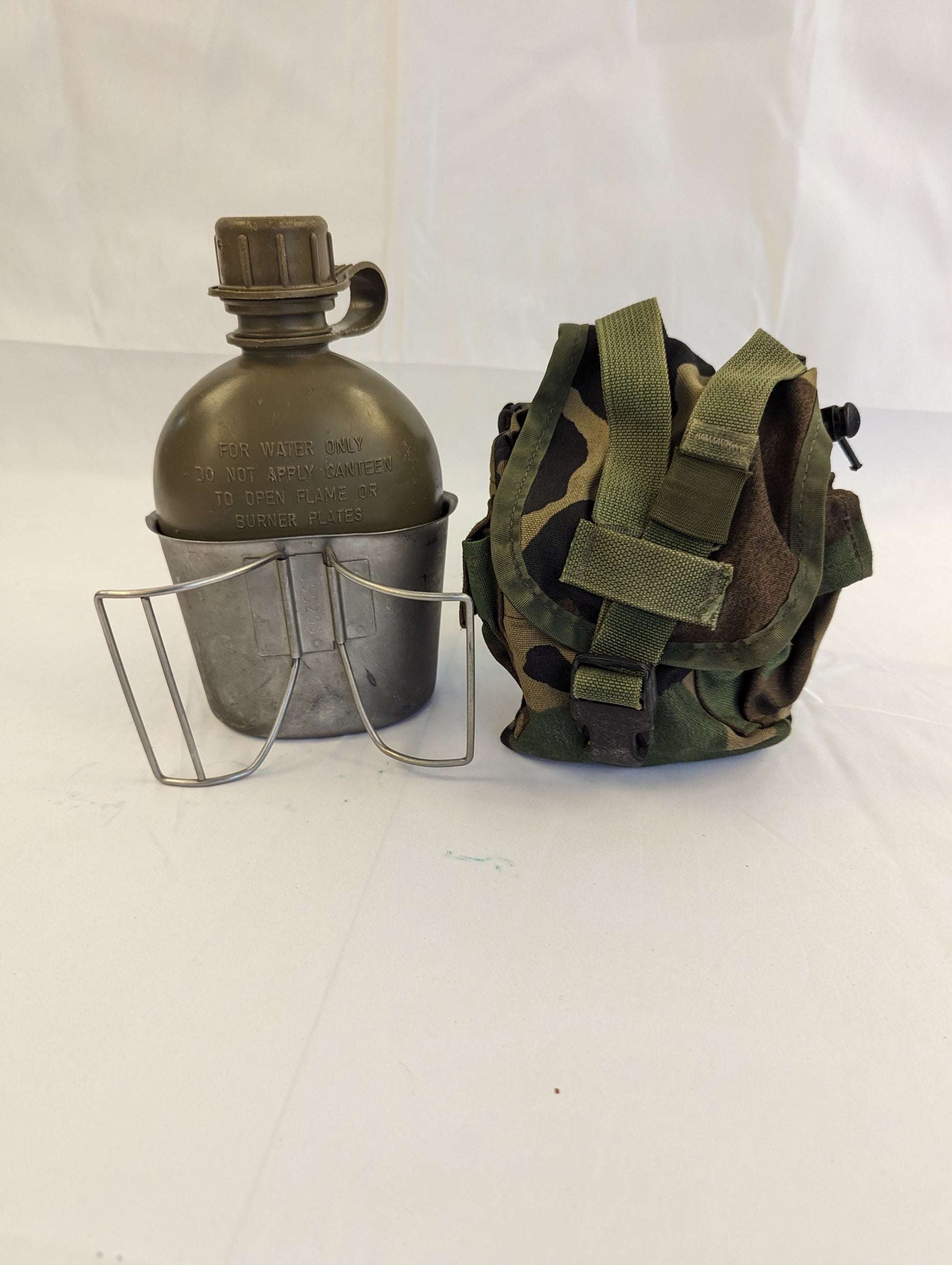 A studio shot of the three components of a U.S. Military 1-quart canteen set: an olive green canteen, a metal canteen cup, and a Woodland camouflage fabric pouch, displayed on a white surface.
