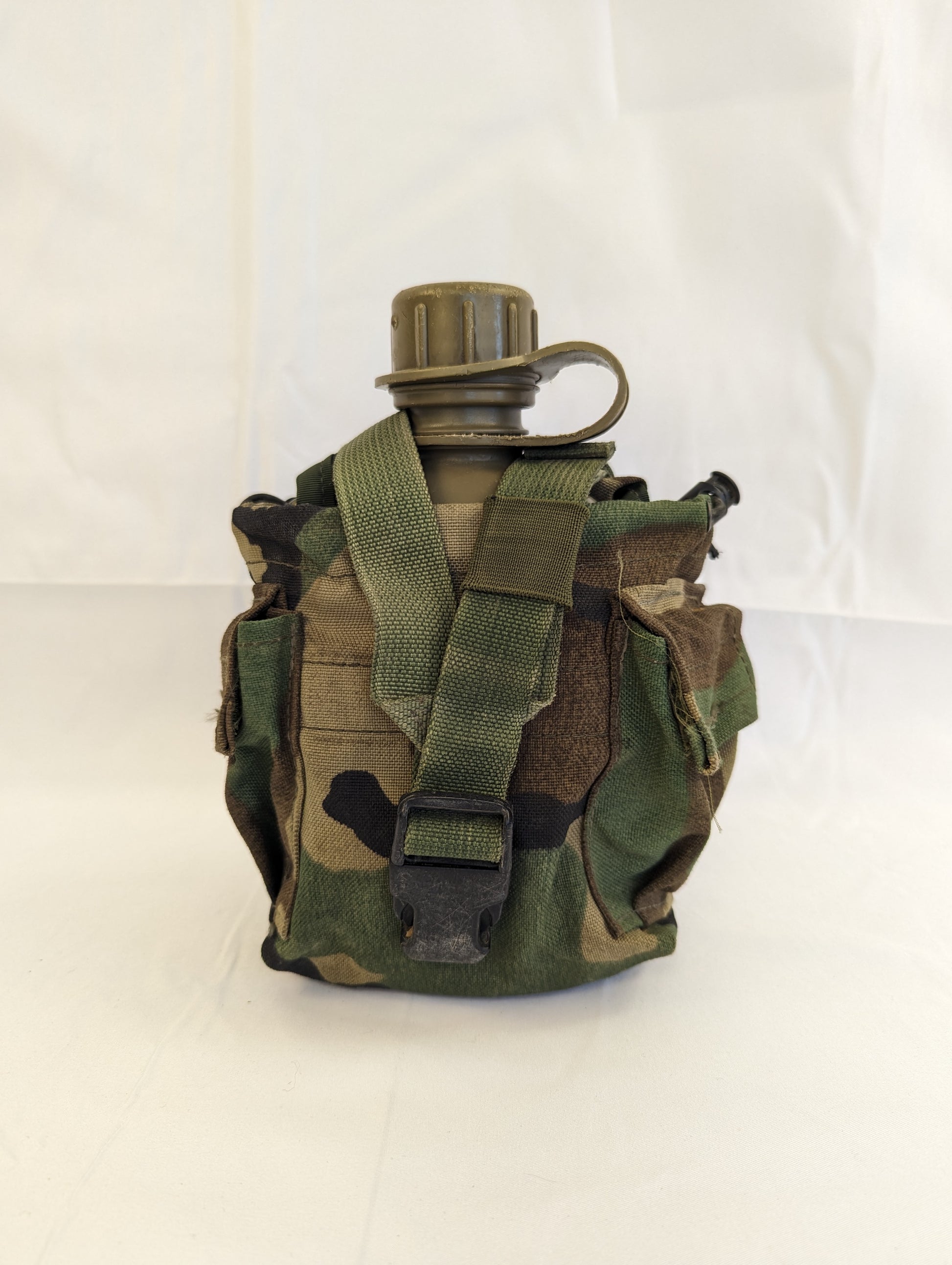 A U.S. Military Surplus 1-quart canteen fully contained within its Woodland camouflage pouch, featuring a black quick-release buckle and the olive green canteen cap visible at the top.