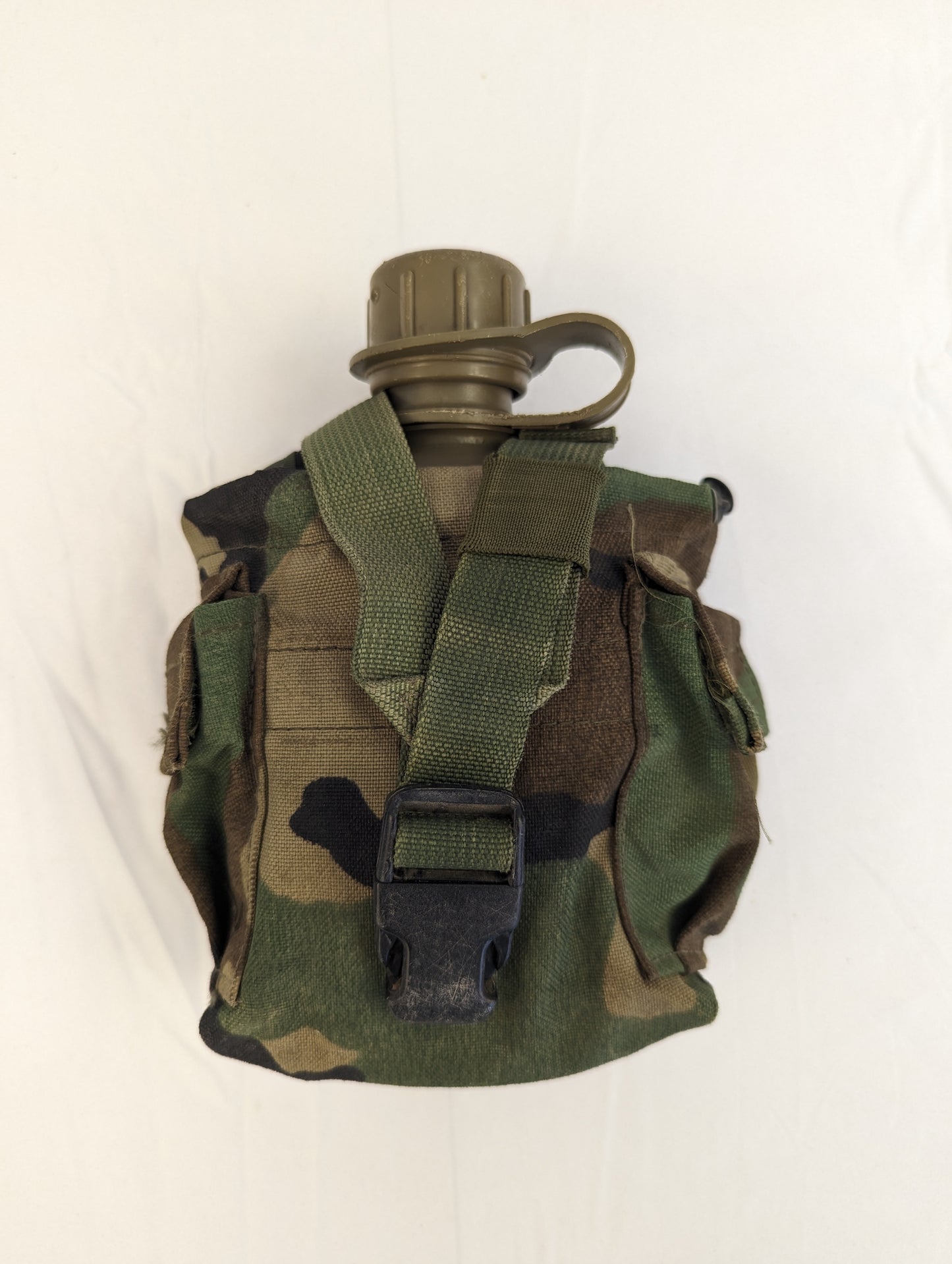 A U.S. Military 1-quart canteen in olive green, stored inside a Woodland camouflage pouch, with the black quick-release buckle and cap visible, displayed on a white background.