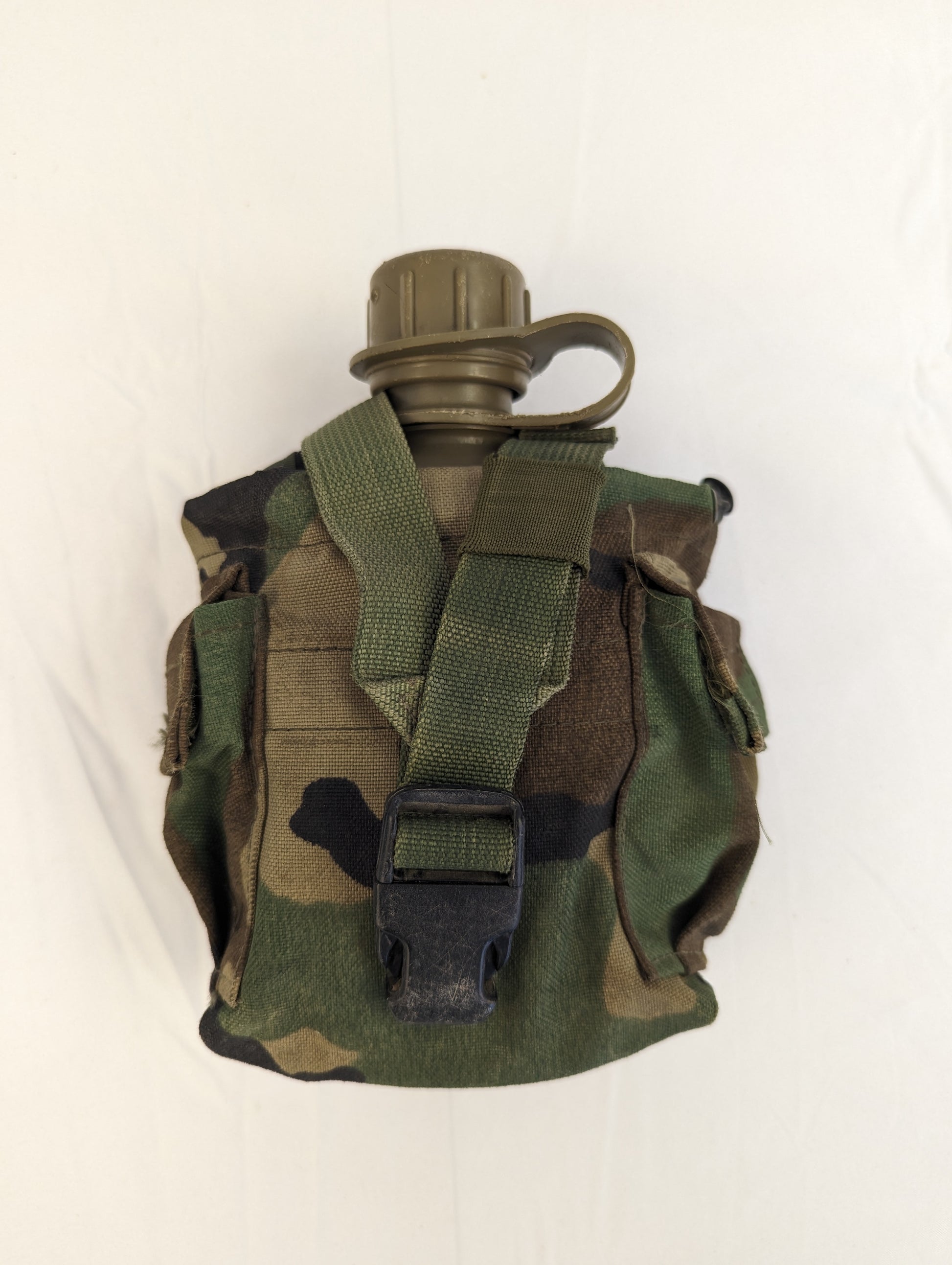 A U.S. Military 1-quart canteen in olive green, stored inside a Woodland camouflage pouch, with the black quick-release buckle and cap visible, displayed on a white background.