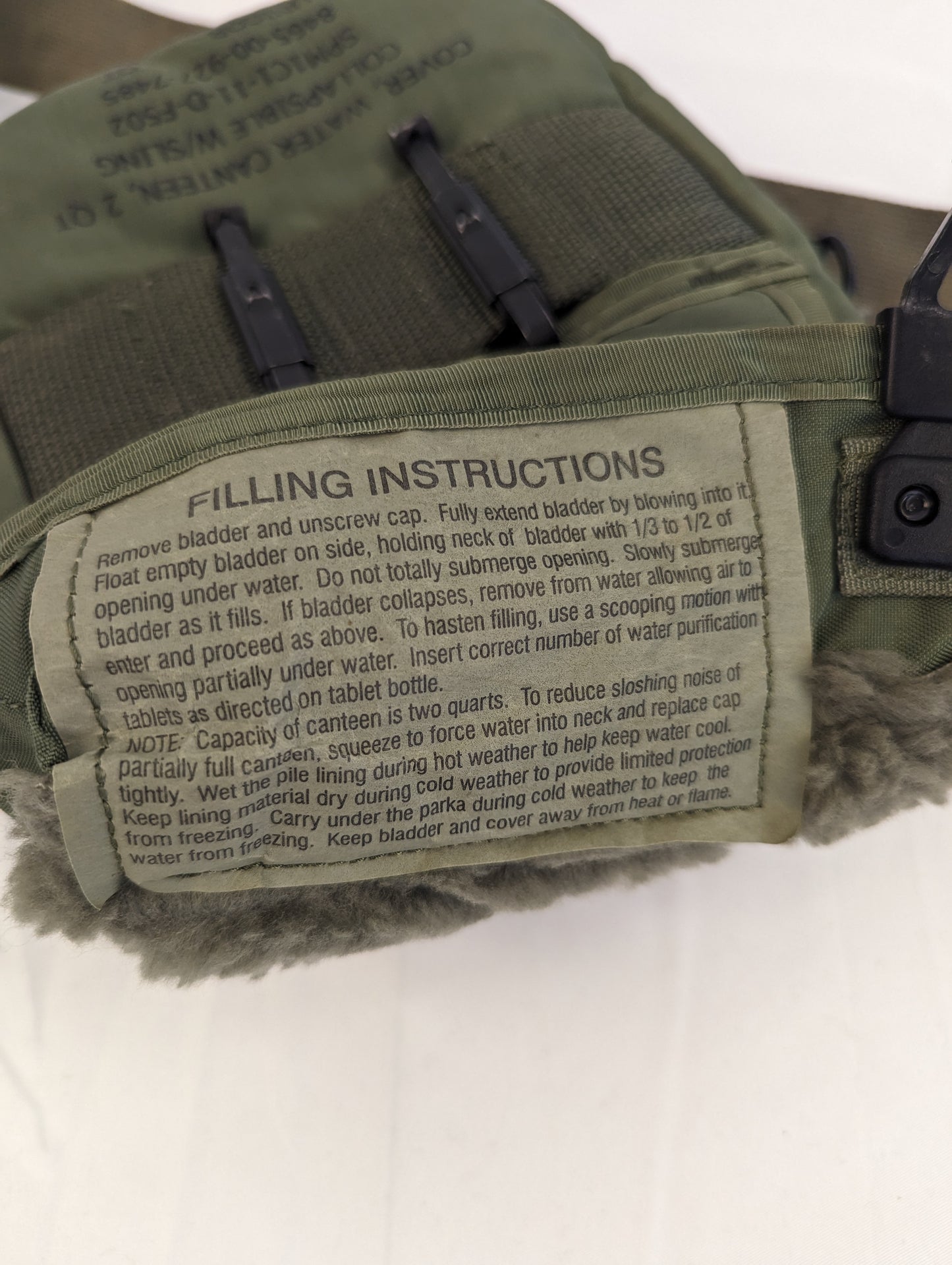 A close-up photograph of the 'FILLING INSTRUCTIONS' label sewn onto the U.S. Military 2-quart canteen cover, providing detailed steps for filling the bladder and wetting the pile lining for cooling.