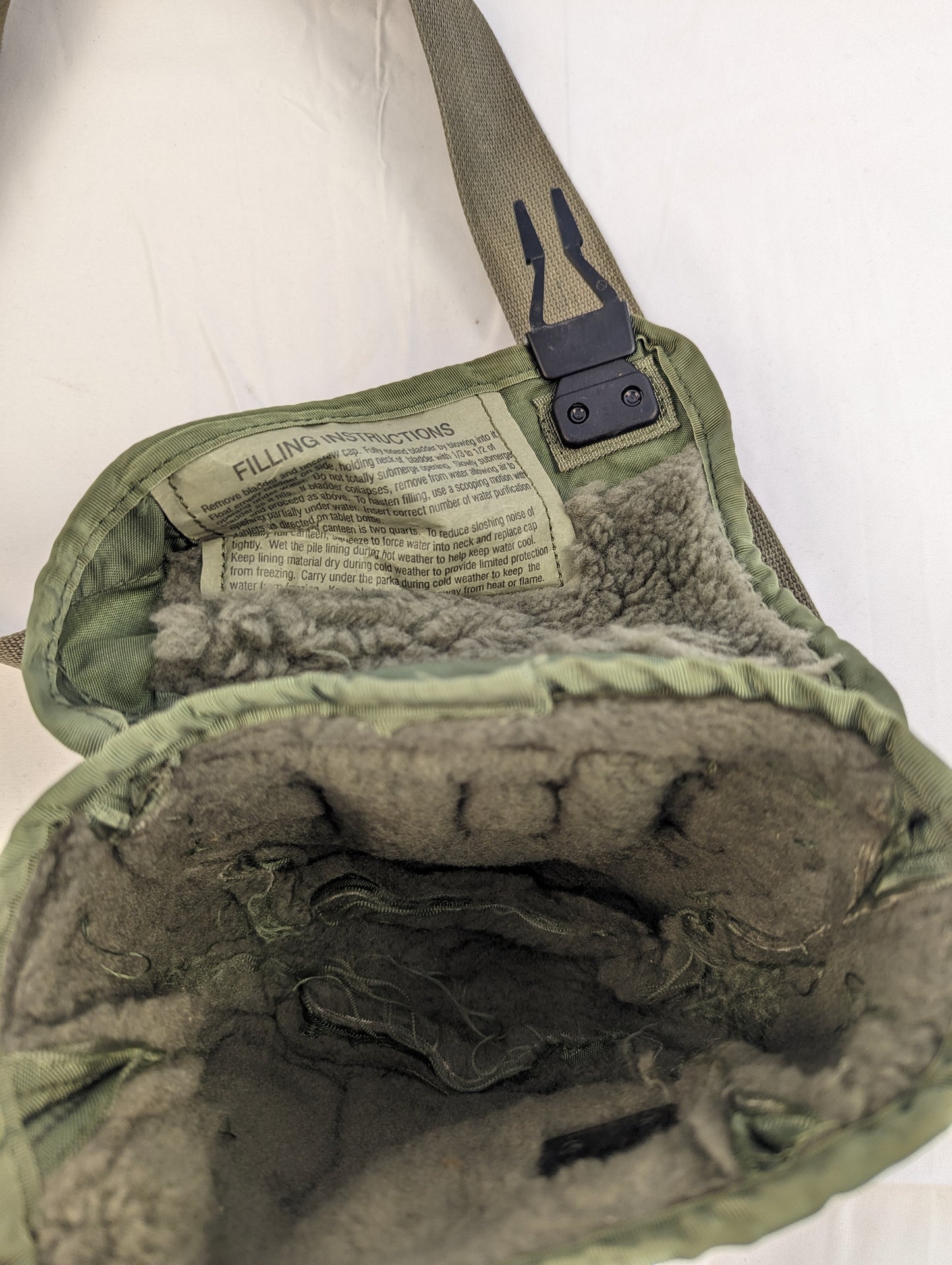 A close-up view into the open interior of a U.S. Military 2-quart canteen cover in olive green, displaying the synthetic fur lining and the 'FILLING INSTRUCTIONS' label sewn onto the flap.