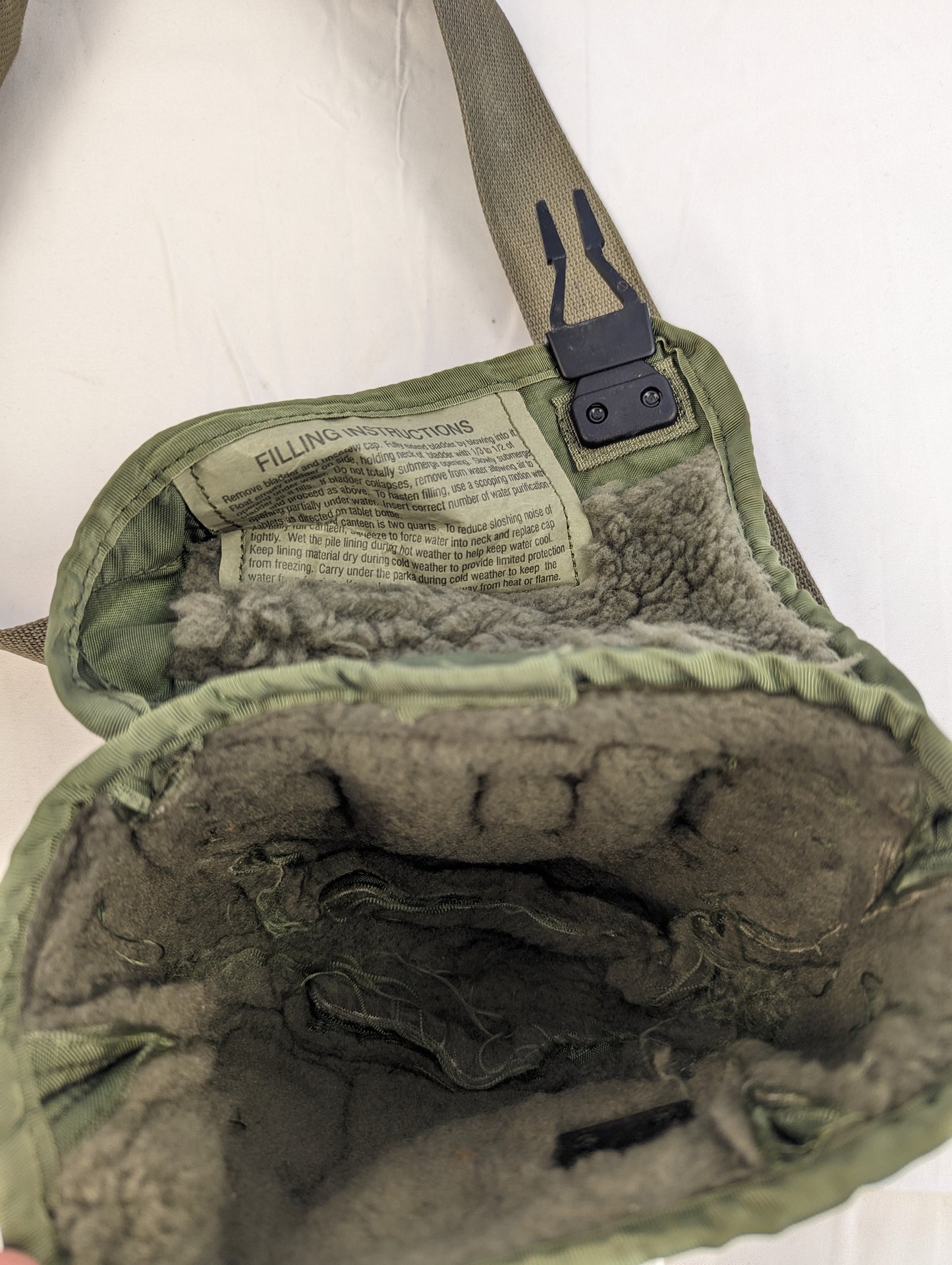 A close-up view into the open interior of a U.S. Military 2-quart canteen cover in olive green, displaying the synthetic fur lining and the 'FILLING INSTRUCTIONS' label sewn onto the flap.