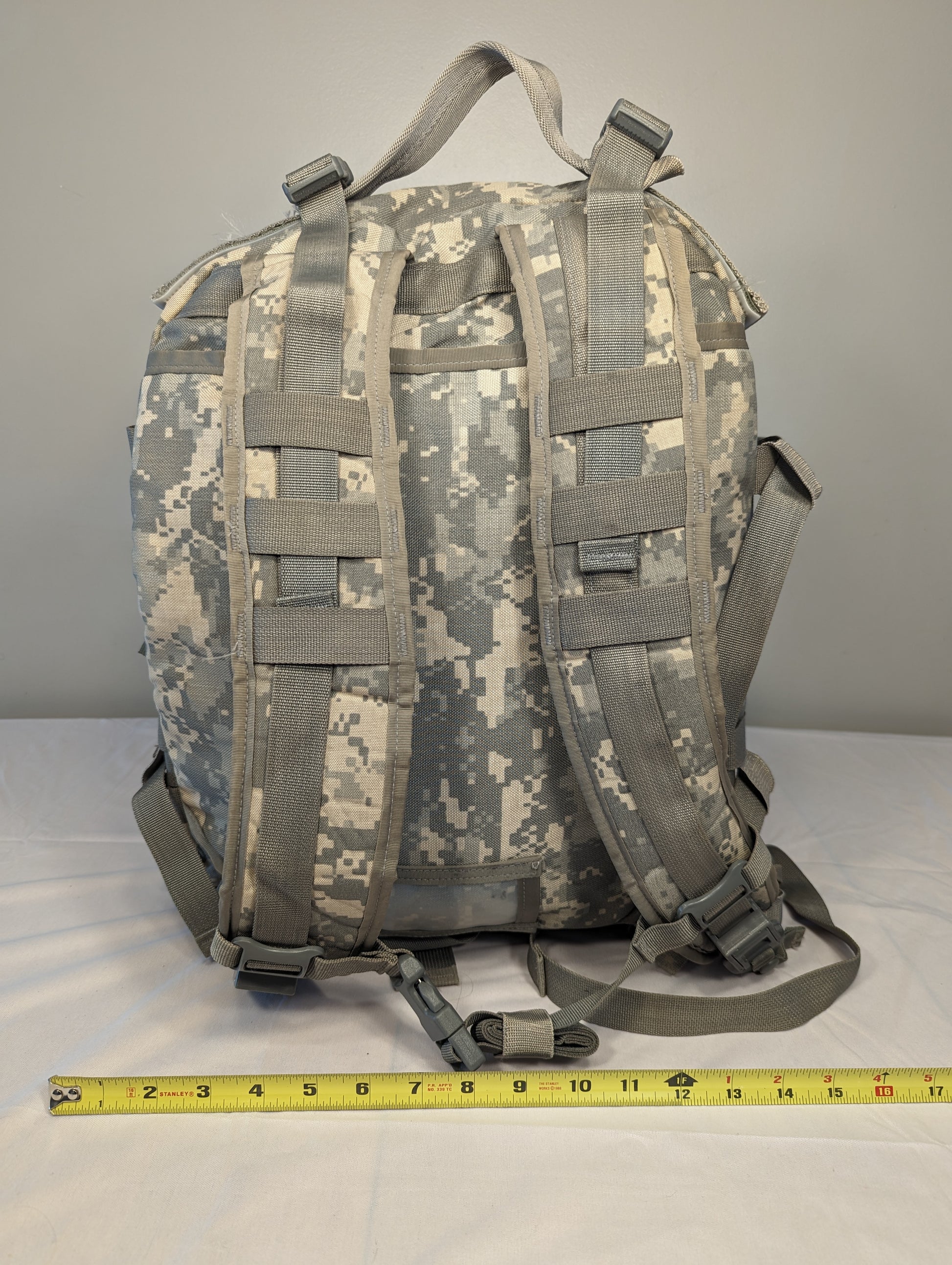A rear view of a U.S. Military Surplus MOLLE II 3-Day Assault Pack in ACU digital camouflage, standing upright, highlighting the padded shoulder straps, suspension webbing, and a tape measure at the base showing the width.