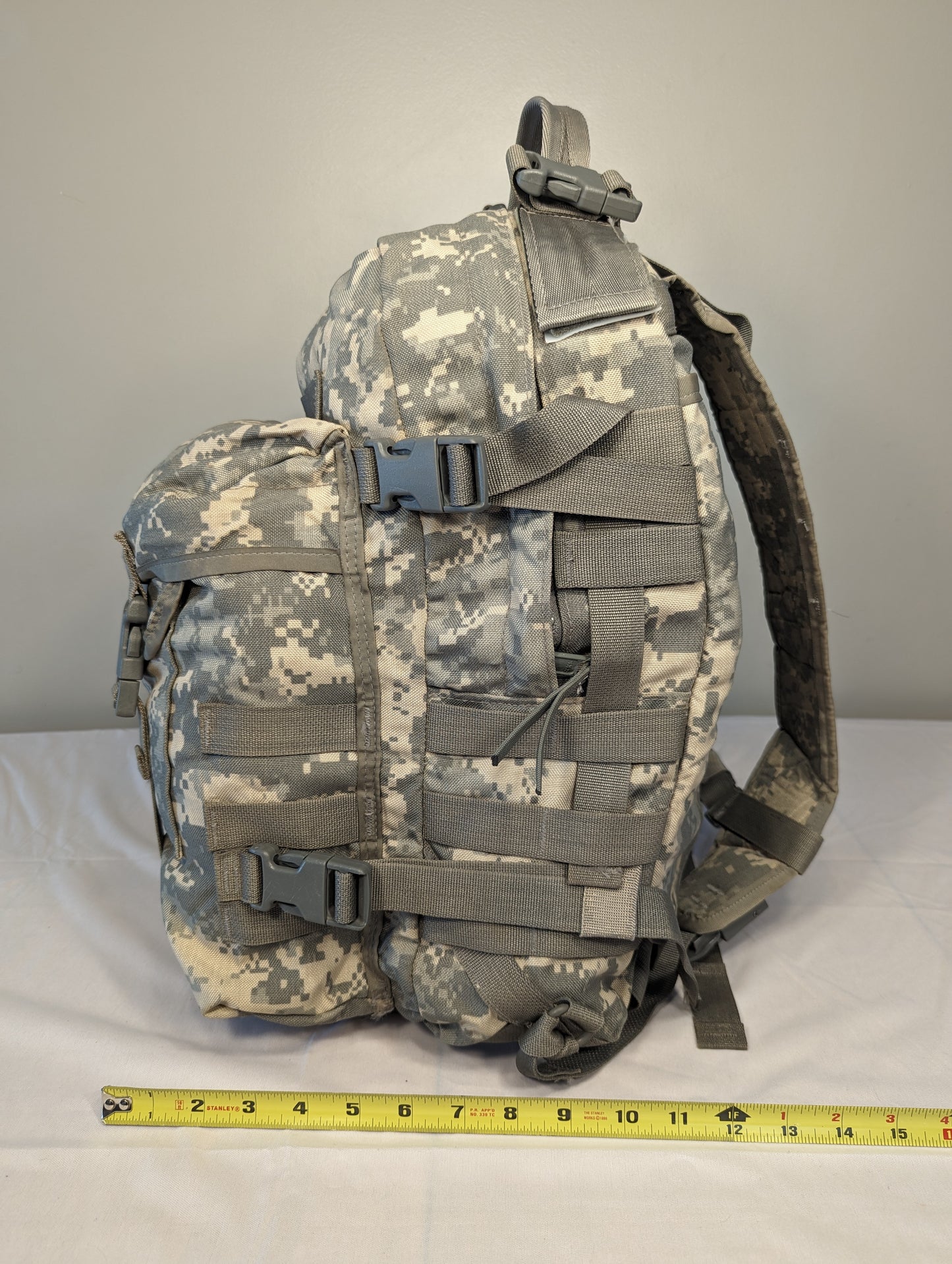 A U.S. Military Surplus MOLLE II 3-Day Assault Pack in ACU digital camouflage, standing upright and featuring compression straps, quick-release buckles, and a measuring tape at the base for scale.