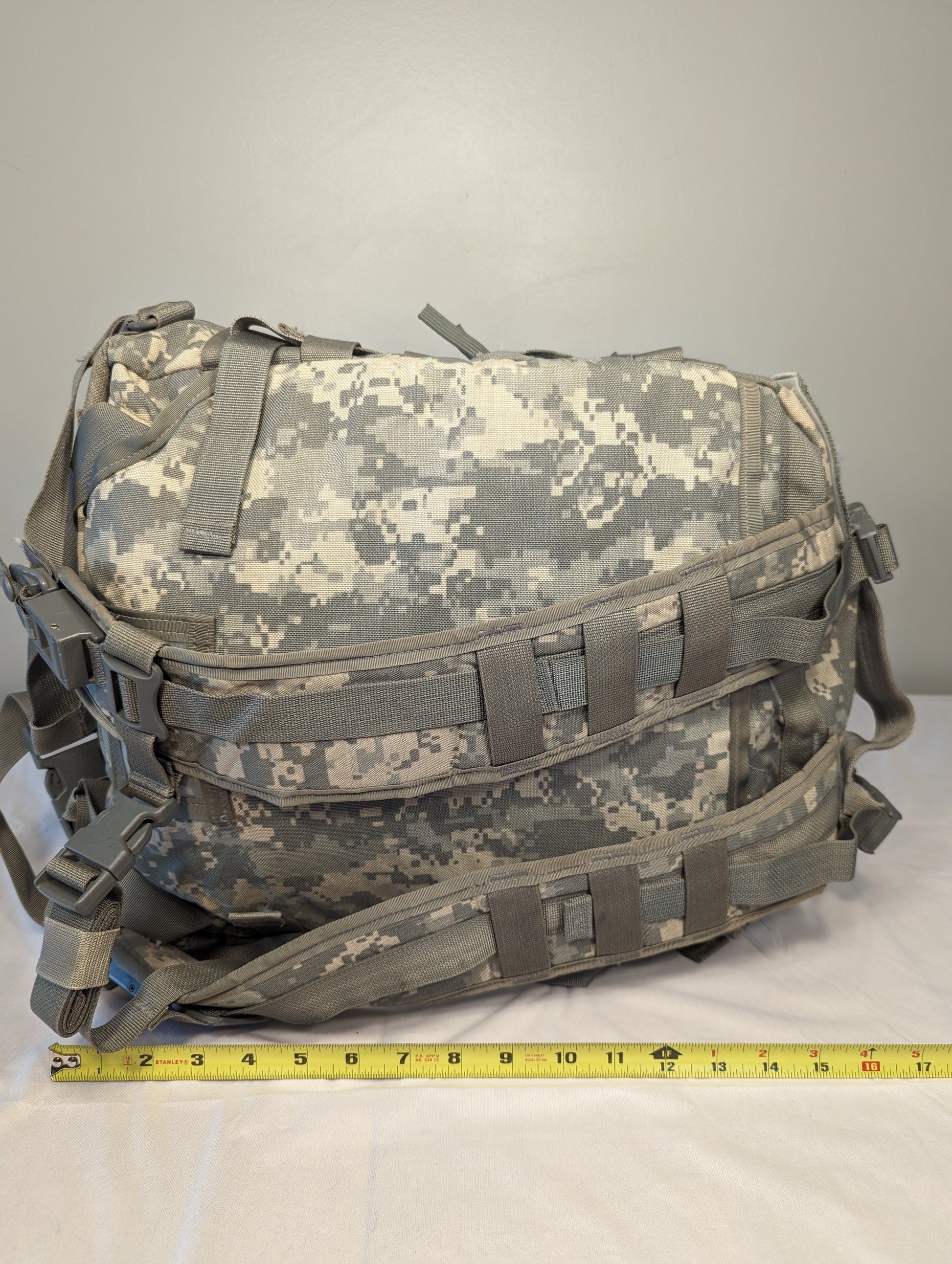 A horizontal view of a U.S. Military Surplus 3-Day Assault Pack in ACU digital camouflage, laid flat with a yellow tape measure demonstrating its width, and secured by MOLLE webbing and compression straps.