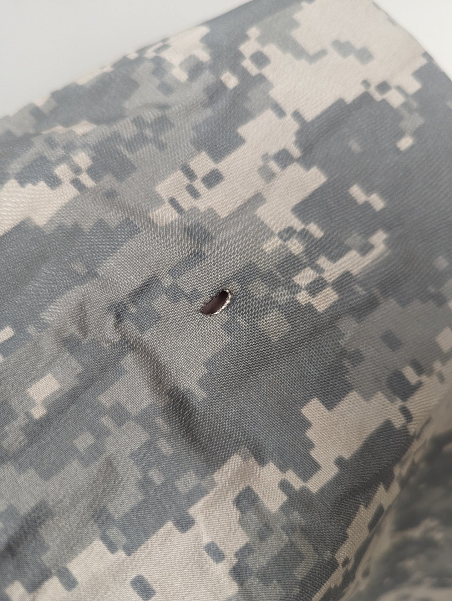 An extreme close-up detail shot of the ACU digital camouflage fabric of the Bivy Cover, clearly showing a burn hole or small tear in the material.