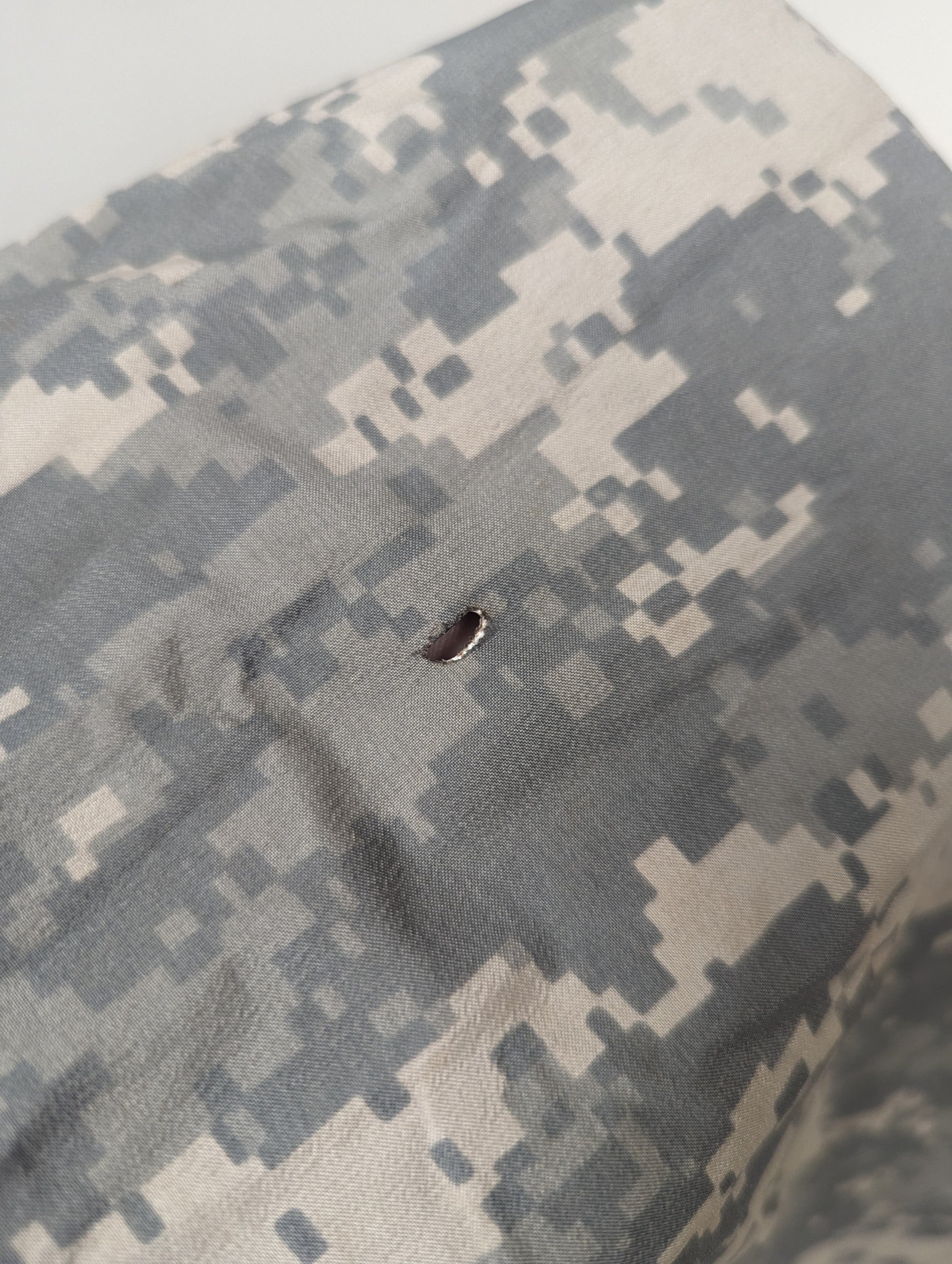 An extreme close-up detail shot of the ACU digital camouflage fabric of the Bivy Cover, clearly showing a burn hole or small tear in the material.