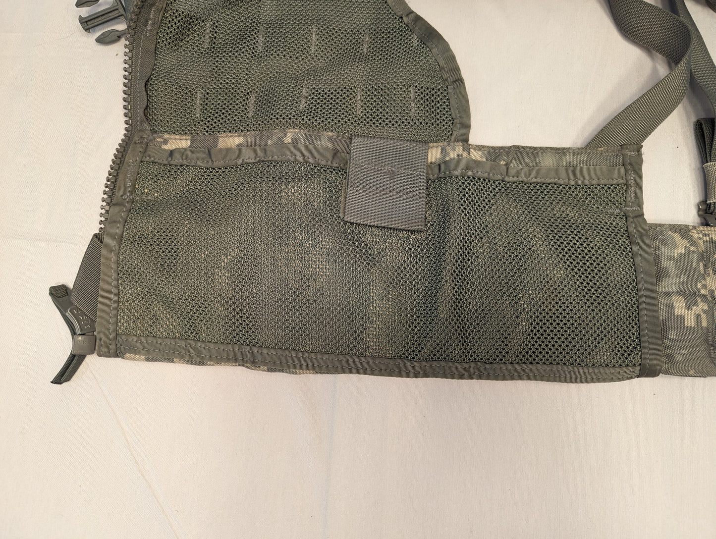 A close-up view of the side panel of the U.S. Military Load Carrier Vest (FLC), highlighting the breathable green mesh body, the extensive horizontal MOLLE webbing, and a compression strap.
