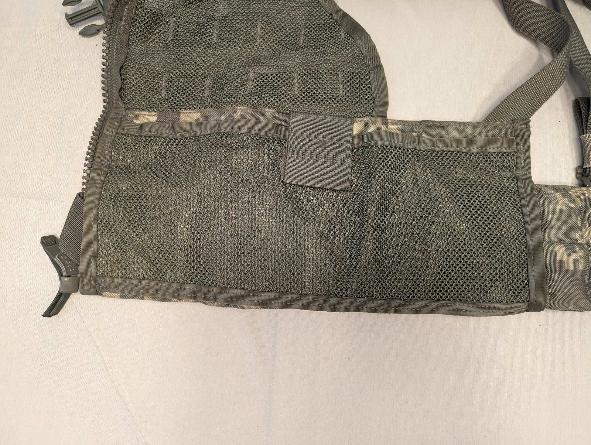 A close-up view of the side panel of the U.S. Military Load Carrier Vest (FLC), highlighting the breathable green mesh body, the extensive horizontal MOLLE webbing, and a compression strap.