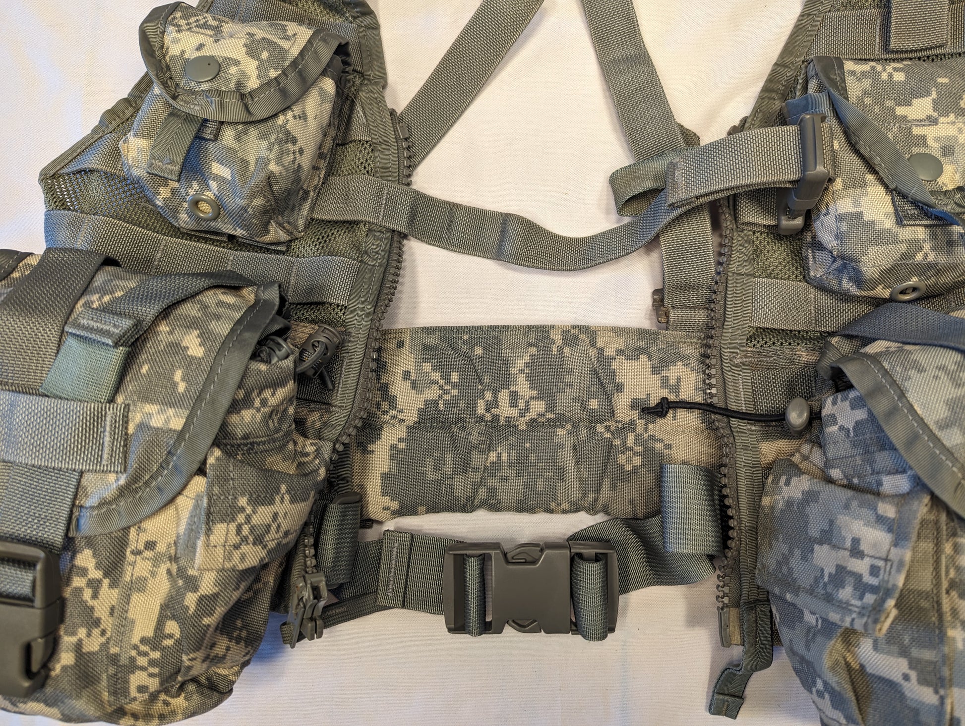 A close-up view of the center panel of a U.S. Military Load Carrier Vest (FLC) in ACU camouflage, highlighting the padded waist belt, the large quick-release buckle, and the attached magazine pouches on either side.