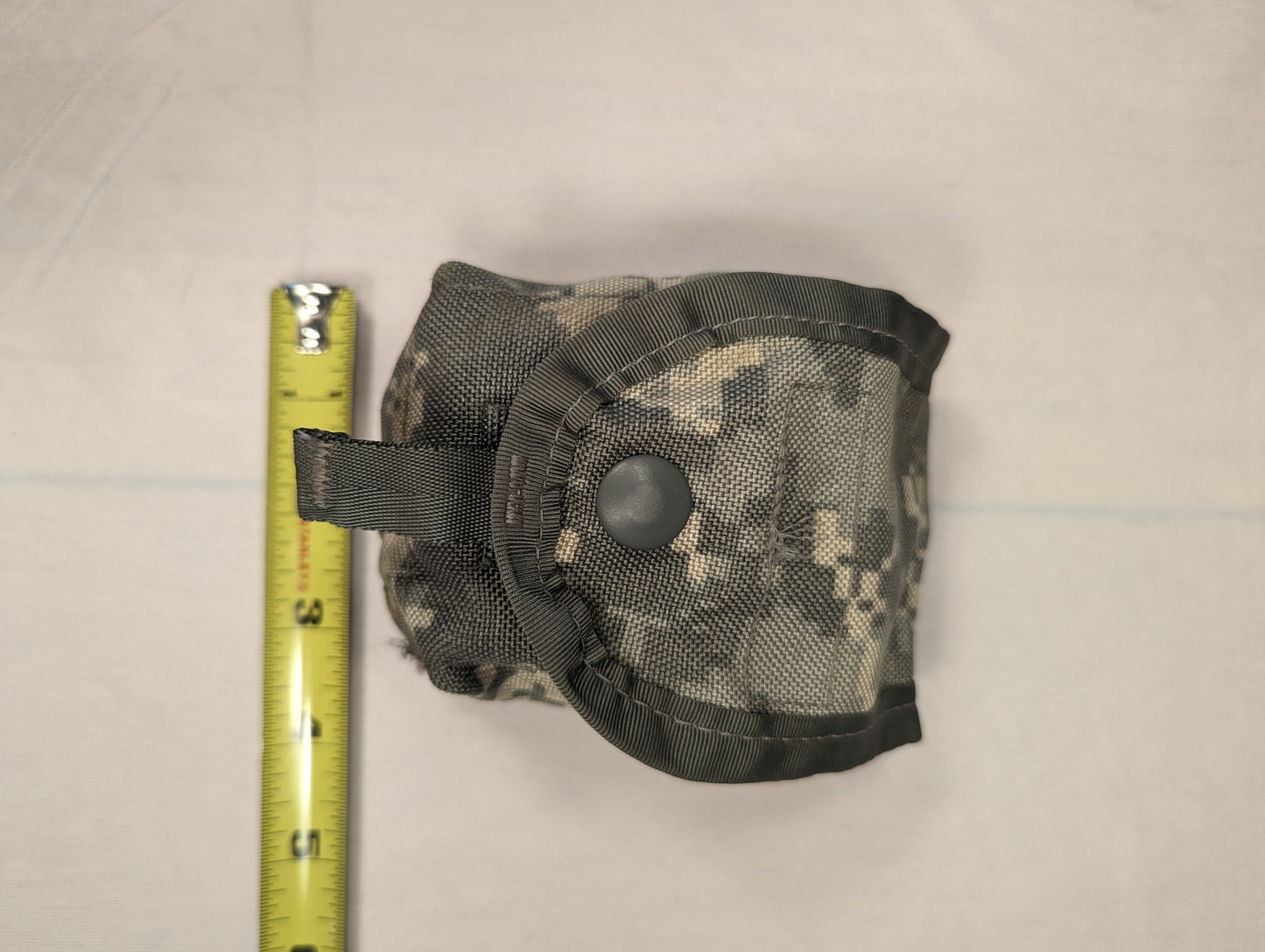 A tightly rolled U.S. Military Surplus Hand Grenade Pouch in ACU digital camouflage, secured with a large black snap button flap, with a yellow tape measure placed alongside to demonstrate its compact 3-inch size.