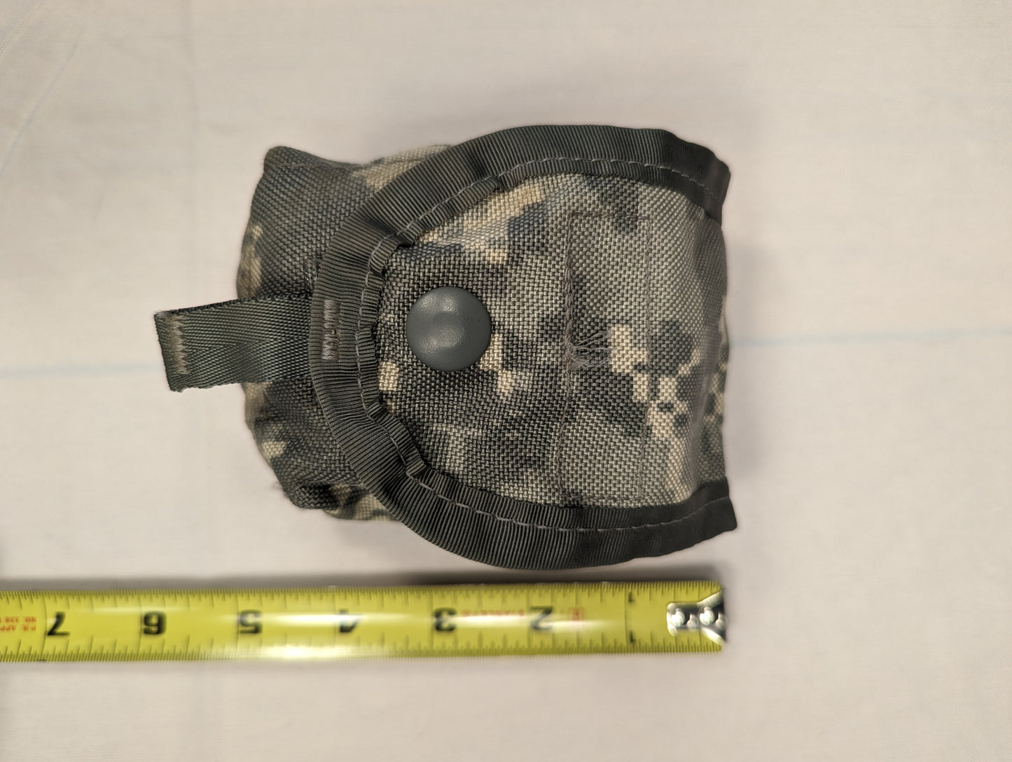 A tightly rolled U.S. Military Surplus Hand Grenade Pouch in ACU digital camouflage, secured with a large snap button closure, with a yellow tape measure below showing the width of approximately 3 to 4 inches.