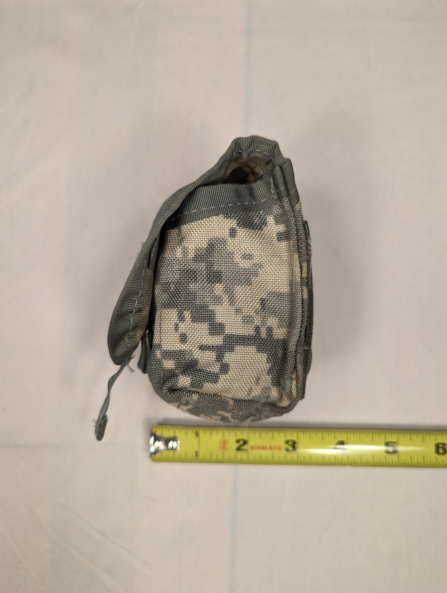 A single U.S. Military Surplus Hand Grenade Pouch in ACU digital camouflage, standing upright with the top flap open, and a yellow tape measure placed at the base demonstrating its compact width.