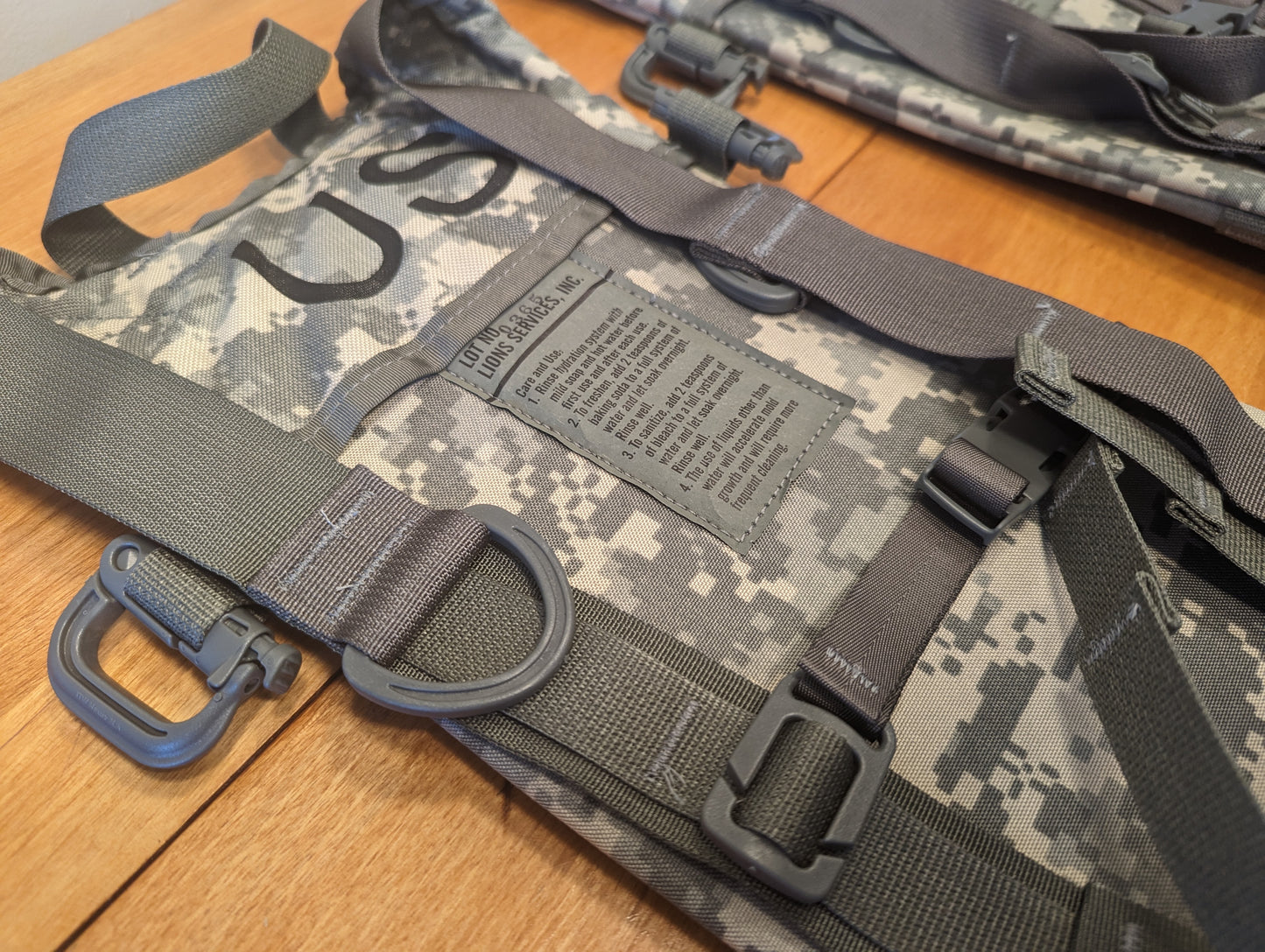 A close-up detail shot of the shoulder strap assembly on a U.S. Military ACU Hydration Carrier, highlighting the large black D-ring, adjustment buckles, and the stenciled 'US' mark.