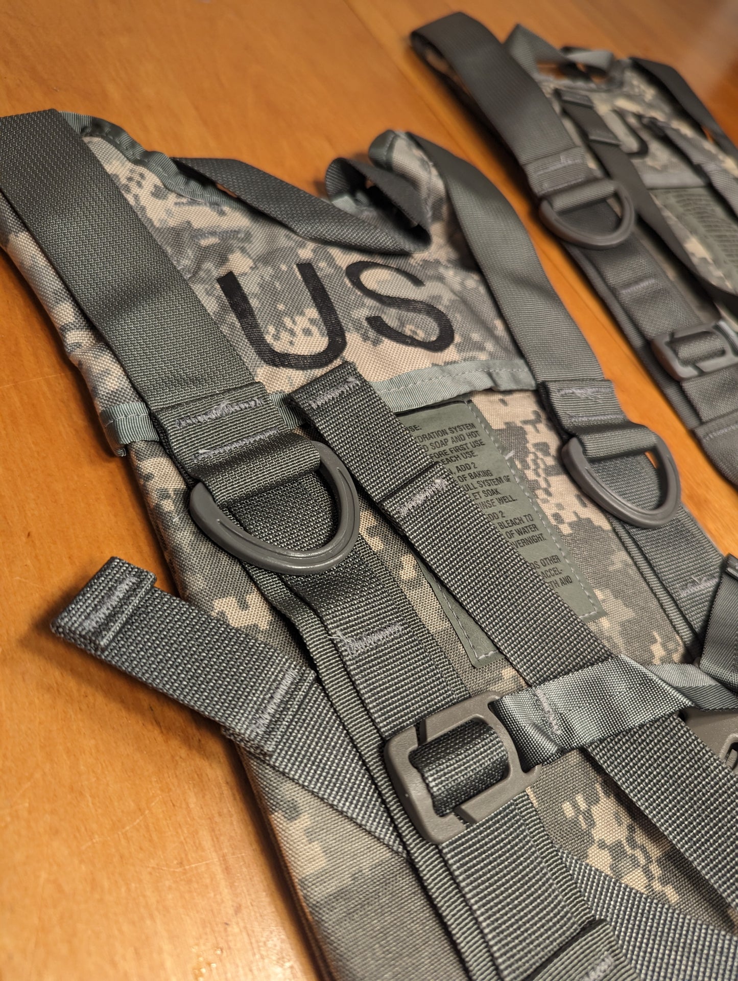 A close-up detail shot of the black heavy-duty webbing and D-ring attachment system of a U.S. Military ACU Hydration Carrier, with the 'US' marking visible on the camouflage fabric.