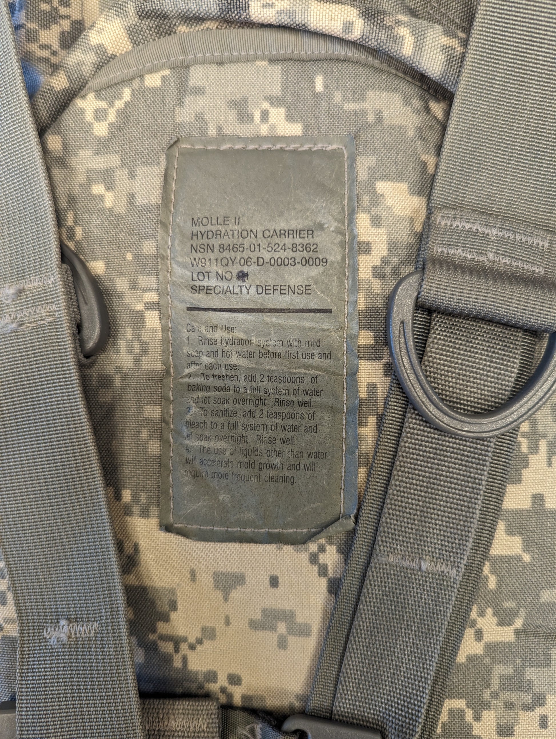 A close-up photograph of the U.S. Military issue tag sewn into the ACU camouflage Hydration Carrier, displaying 'MOLLE II HYDRATION CARRIER', the NSN 8465-01-525-5632, and the manufacturer Specialty Defense.