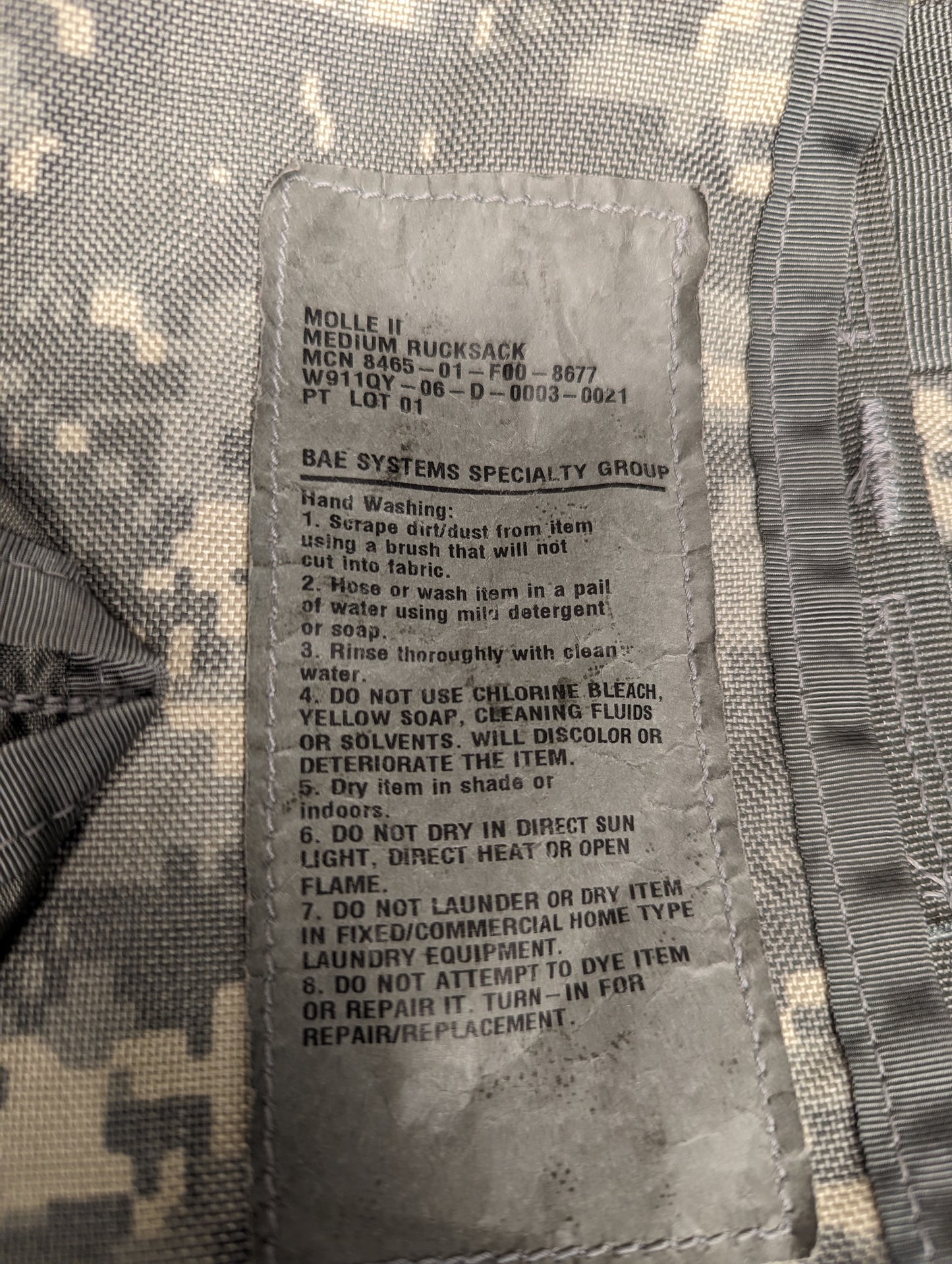 A close-up photograph of the official U.S. Military issue and care tag sewn into the ACU camouflage Medium Field Pack, displaying the product name 'MOLLE MEDIUM PACK', manufacturer BAE Systems, and detailed hand washing instructions.