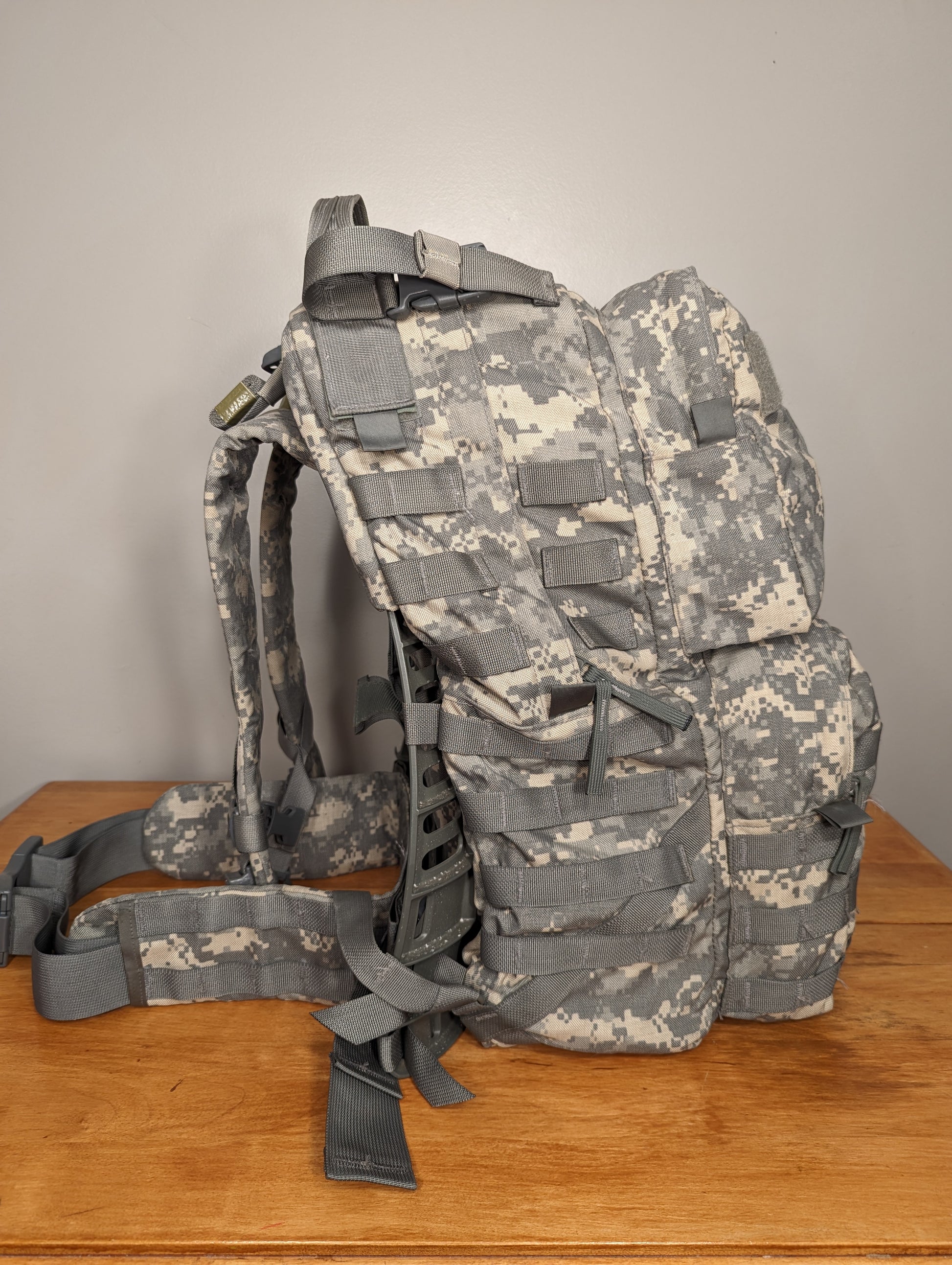 A side view of a U.S. Military Surplus Medium Field Pack in ACU digital camouflage, standing upright on a wooden table, emphasizing the bulky, loaded profile, compression straps, and MOLLE webbing.