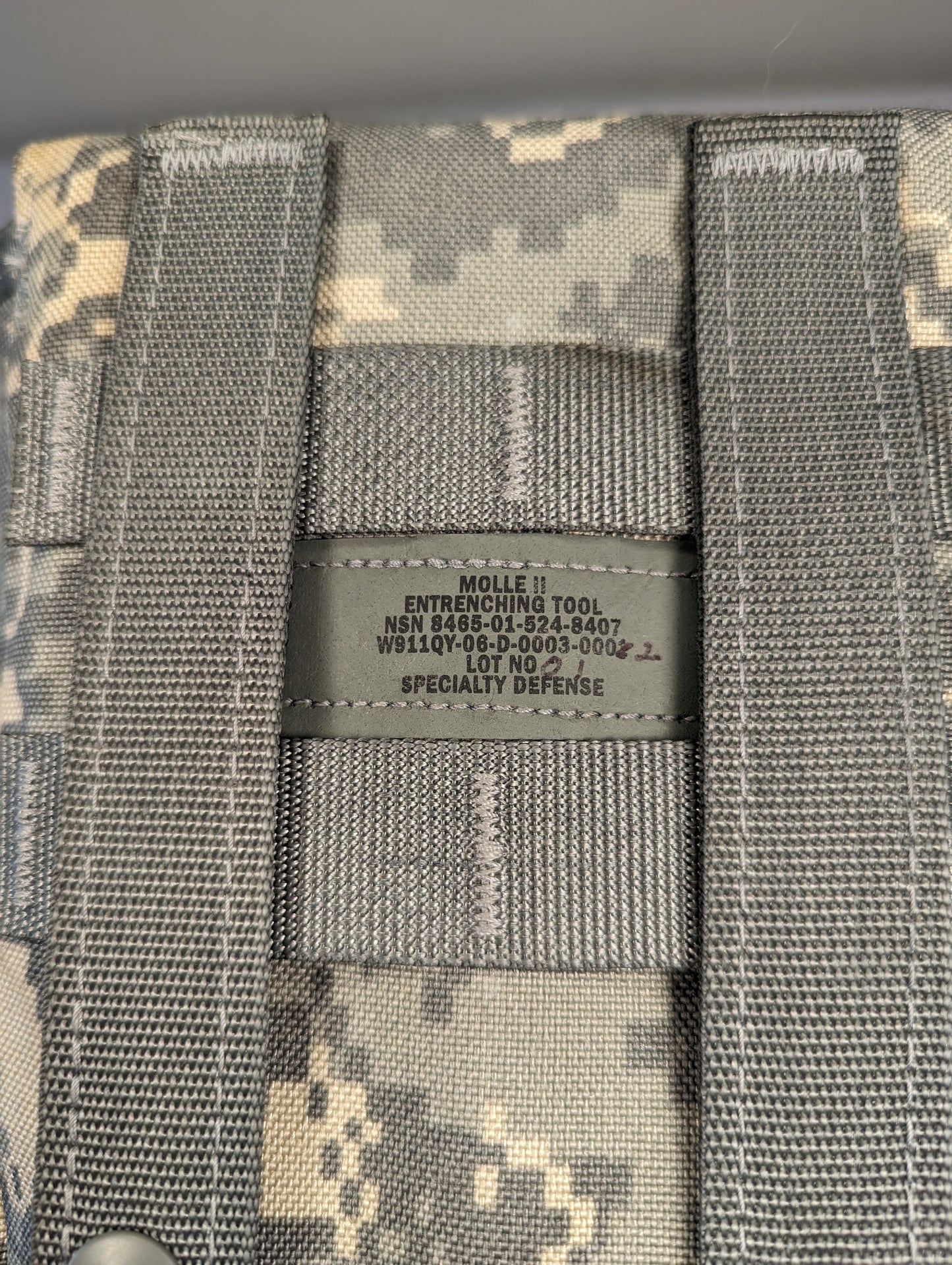A close-up photograph of the official U.S. Military issue tag sewn into the ACU digital camouflage Entrenching Tool Carrier, displaying the product name 'MOLLE II ENTRENCHING TOOL', the NSN 8465-01-524-8407, and manufacturer 'SPECIALTY DEFENSE'.
