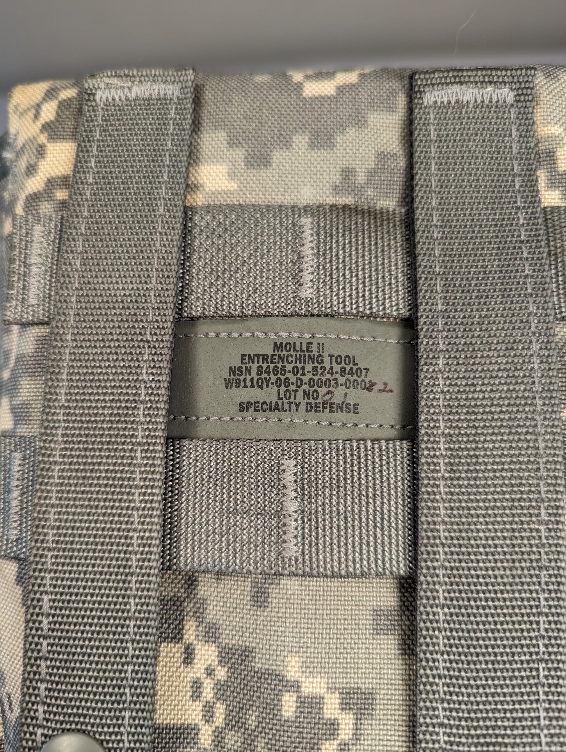 A close-up photograph of the official U.S. Military issue tag sewn onto the ACU digital camouflage Entrenching Tool Carrier, displaying the text 'MOLLE II ENTRENCHING TOOL', the NSN 8465-01-524-8407, and manufacturer 'SPECIALTY DEFENSE'.