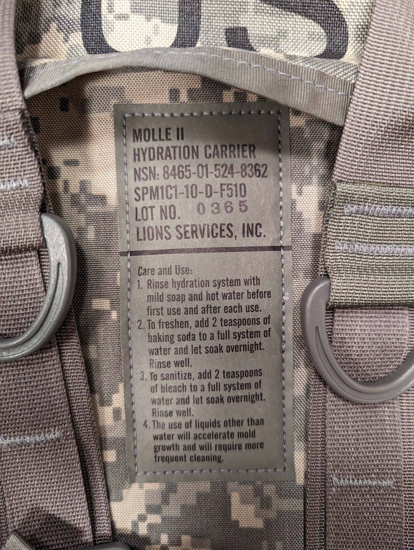 A close-up photograph of the U.S. Military issue and care tag sewn into the ACU camouflage Hydration Carrier, displaying 'MOLLE II HYDRATION CARRIER', NSN 8465-01-524-8252, manufacturer 'Lions Services, Inc.', and detailed cleaning instructions.
