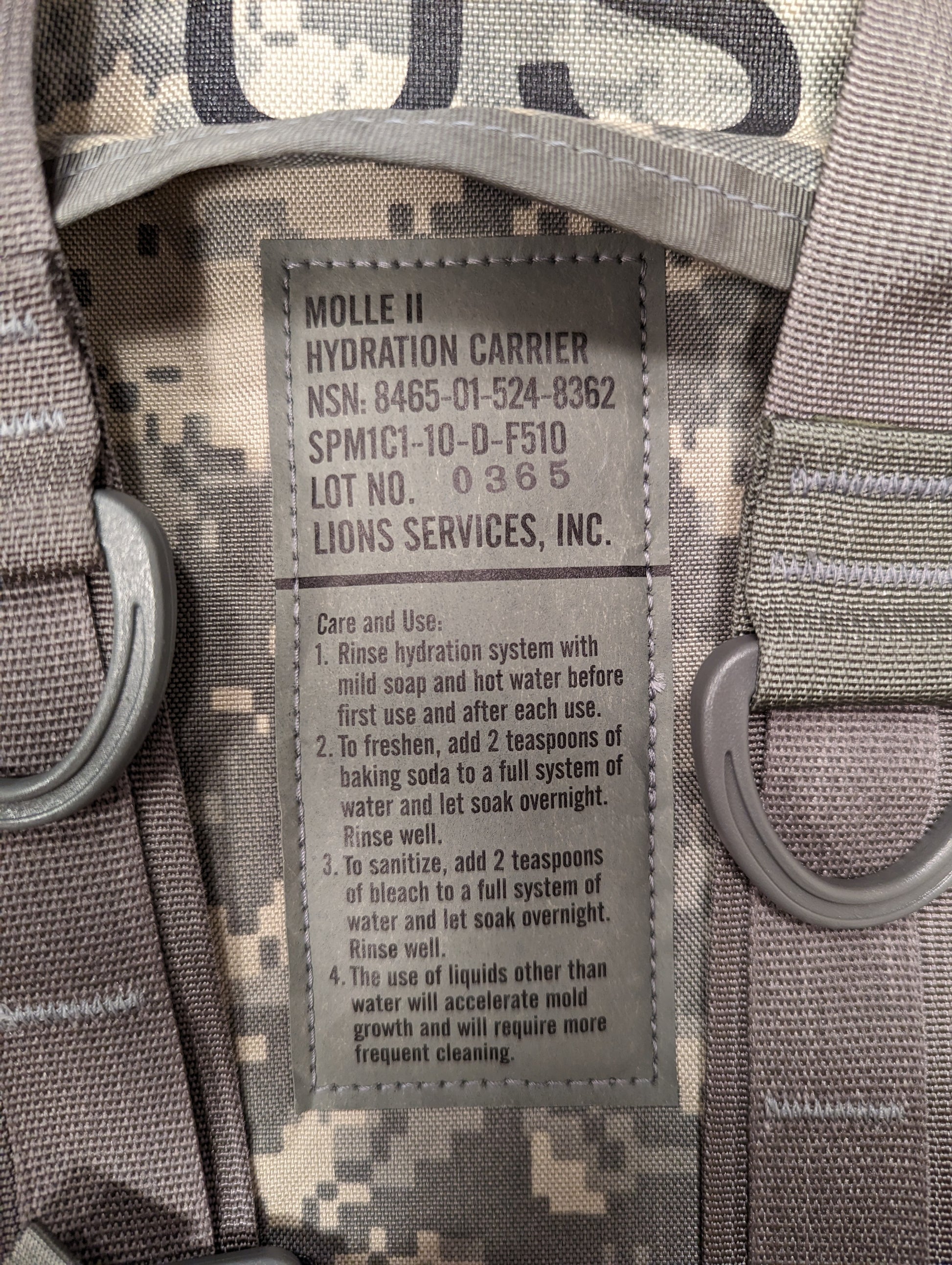 A close-up photograph of the U.S. Military issue and care tag sewn into the ACU camouflage Hydration Carrier, displaying 'MOLLE II HYDRATION CARRIER', NSN 8465-01-524-8252, manufacturer 'Lions Services, Inc.', and detailed cleaning instructions.