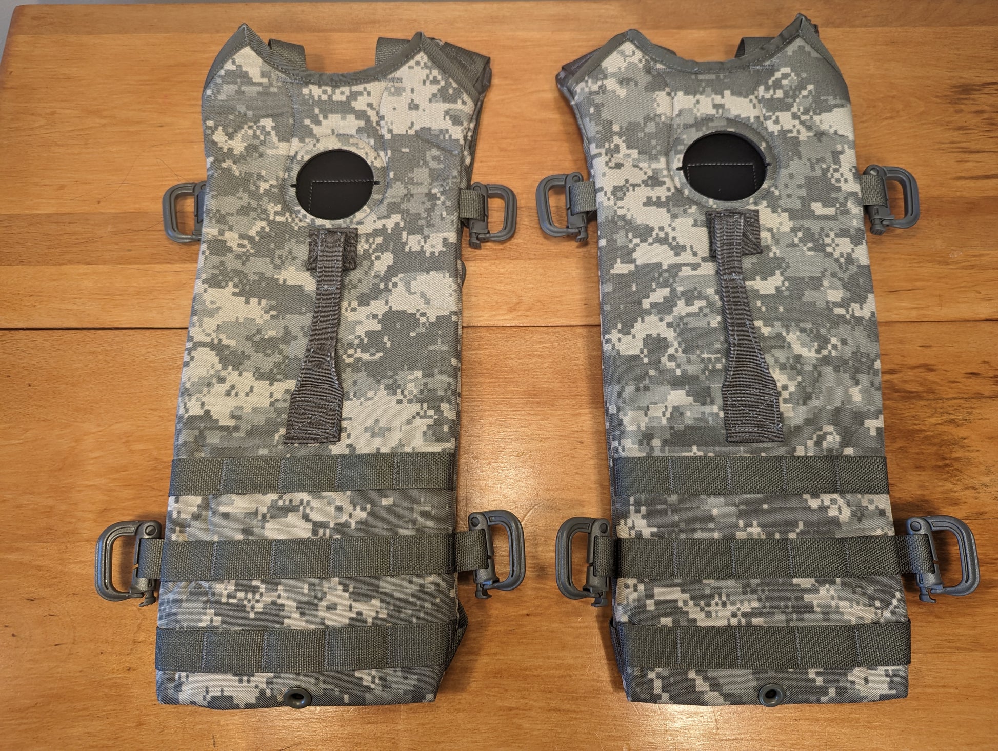 A pair of new U.S. Military Hydration Carrier internal stiffeners in ACU digital camouflage, laid out side-by-side, featuring D-ring attachments and vertical MOLLE webbing on a wooden background.