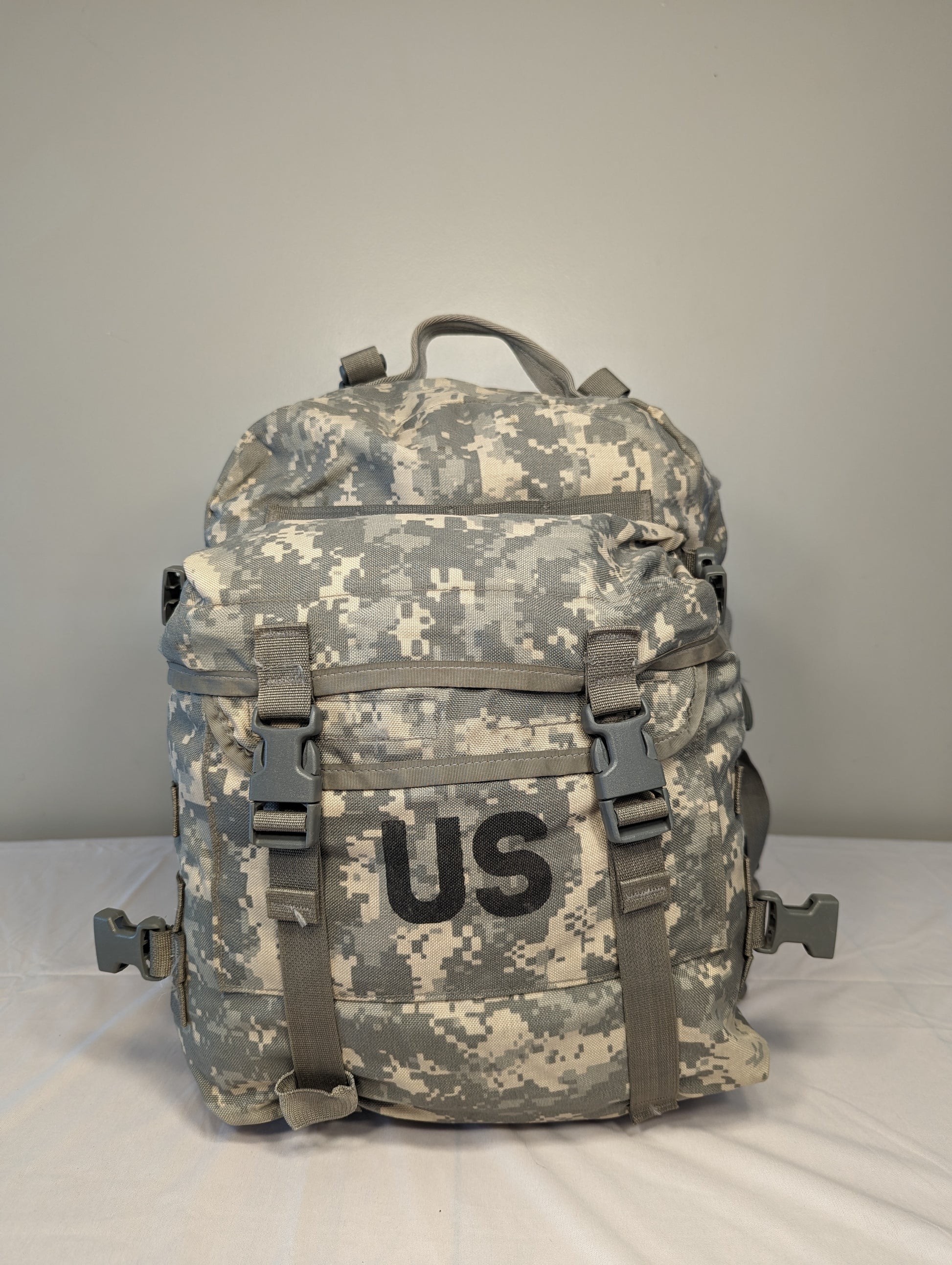 A front view of a U.S. Military Surplus MOLLE II 3-Day Assault Pack in ACU digital camouflage, standing upright, prominently featuring a large stenciled 'US' mark, horizontal MOLLE webbing, and adjustable compression straps.