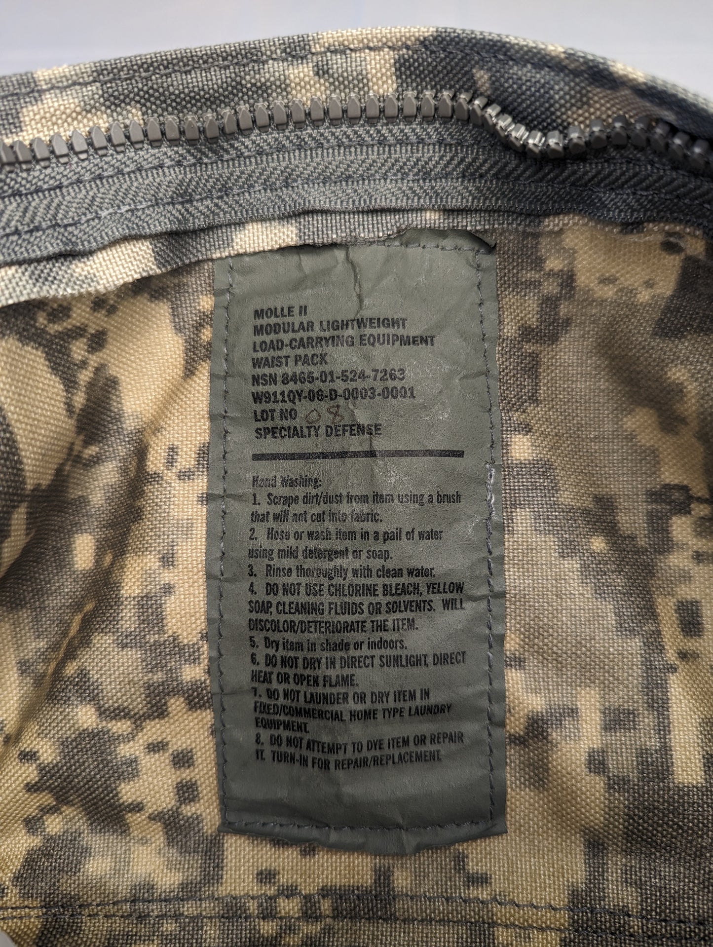 A close-up photograph of the official U.S. Military issue and care tag sewn into the ACU digital camouflage Waist Pack, displaying the product name 'WAIST PACK/CARRIER, E.T./POUCH', NSN 8465-01-524-7262, and detailed usage and care instructions.