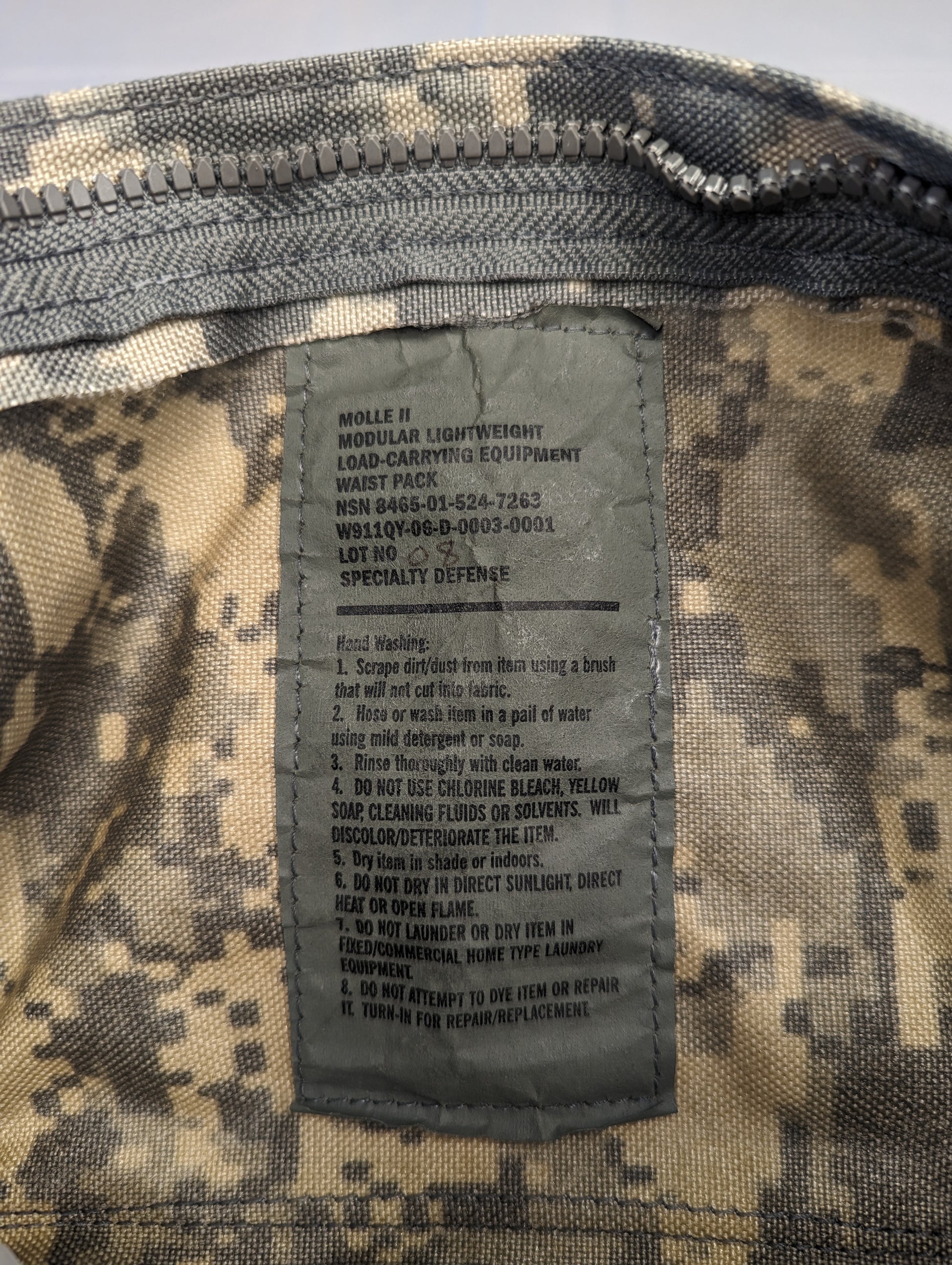 A close-up photograph of the official U.S. Military issue and care tag sewn into the ACU digital camouflage Waist Pack, displaying the product name 'WAIST PACK/CARRIER, E.T./POUCH', NSN 8465-01-524-7262, and detailed usage and care instructions.
